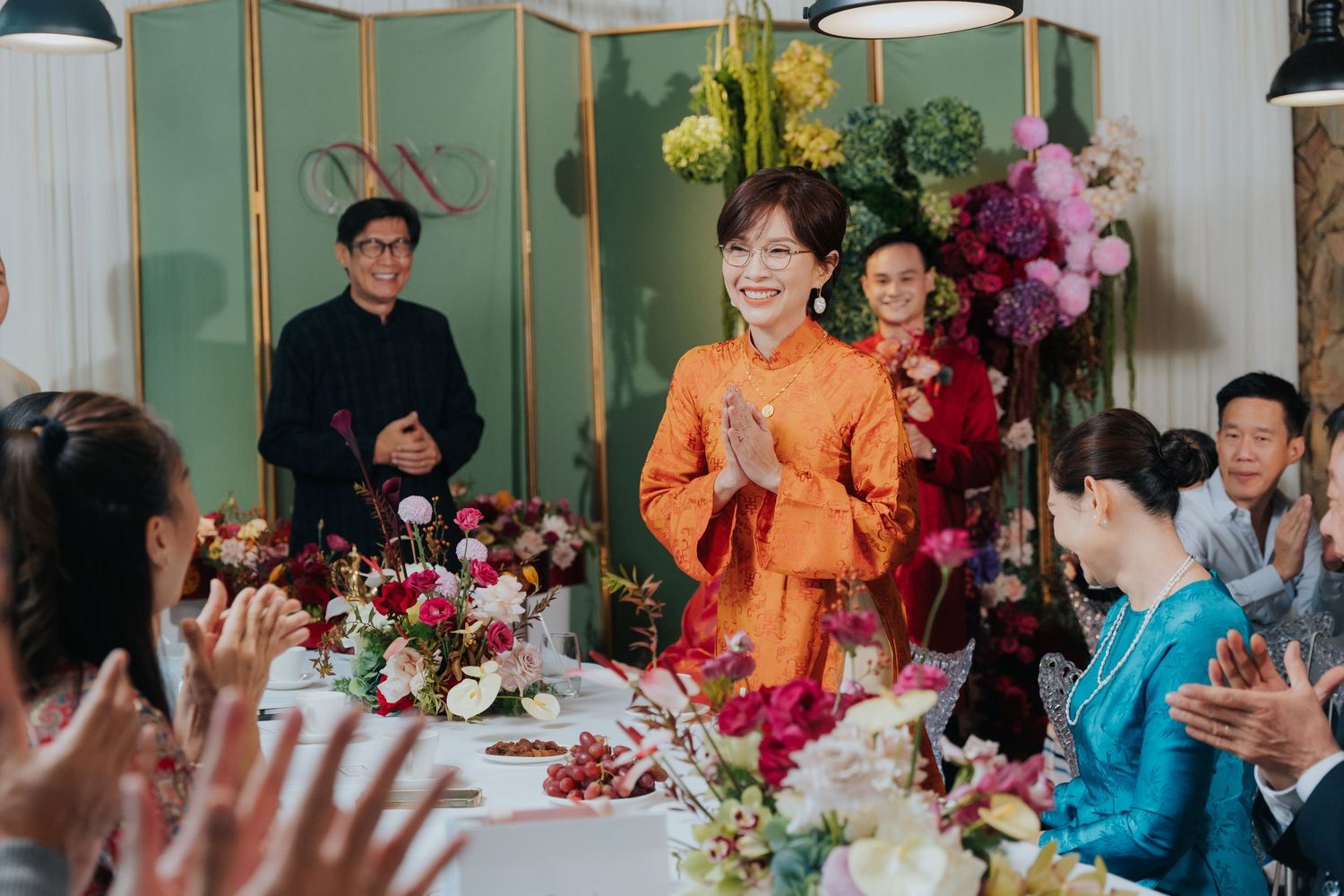 Woman in orange traditional dress greets guests at elegant floral wedding banquet table with colorful decorations.