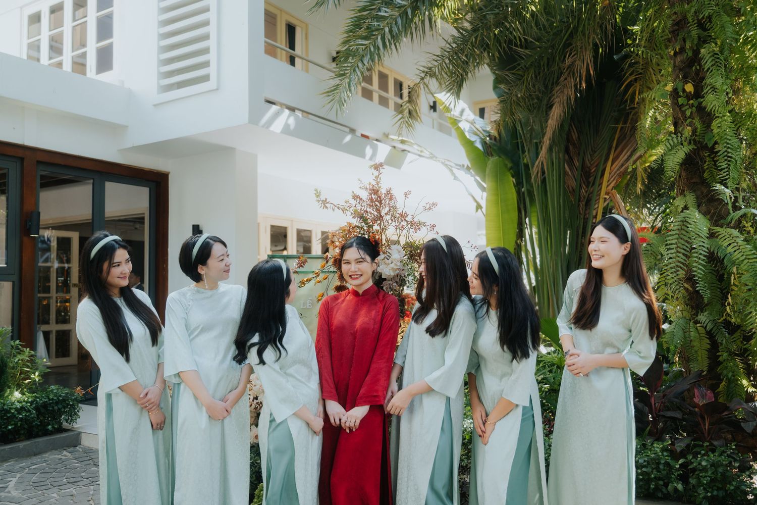 A bride in red ao dai stands with bridesmaids in mint green ao dai gowns in a lush garden setting.