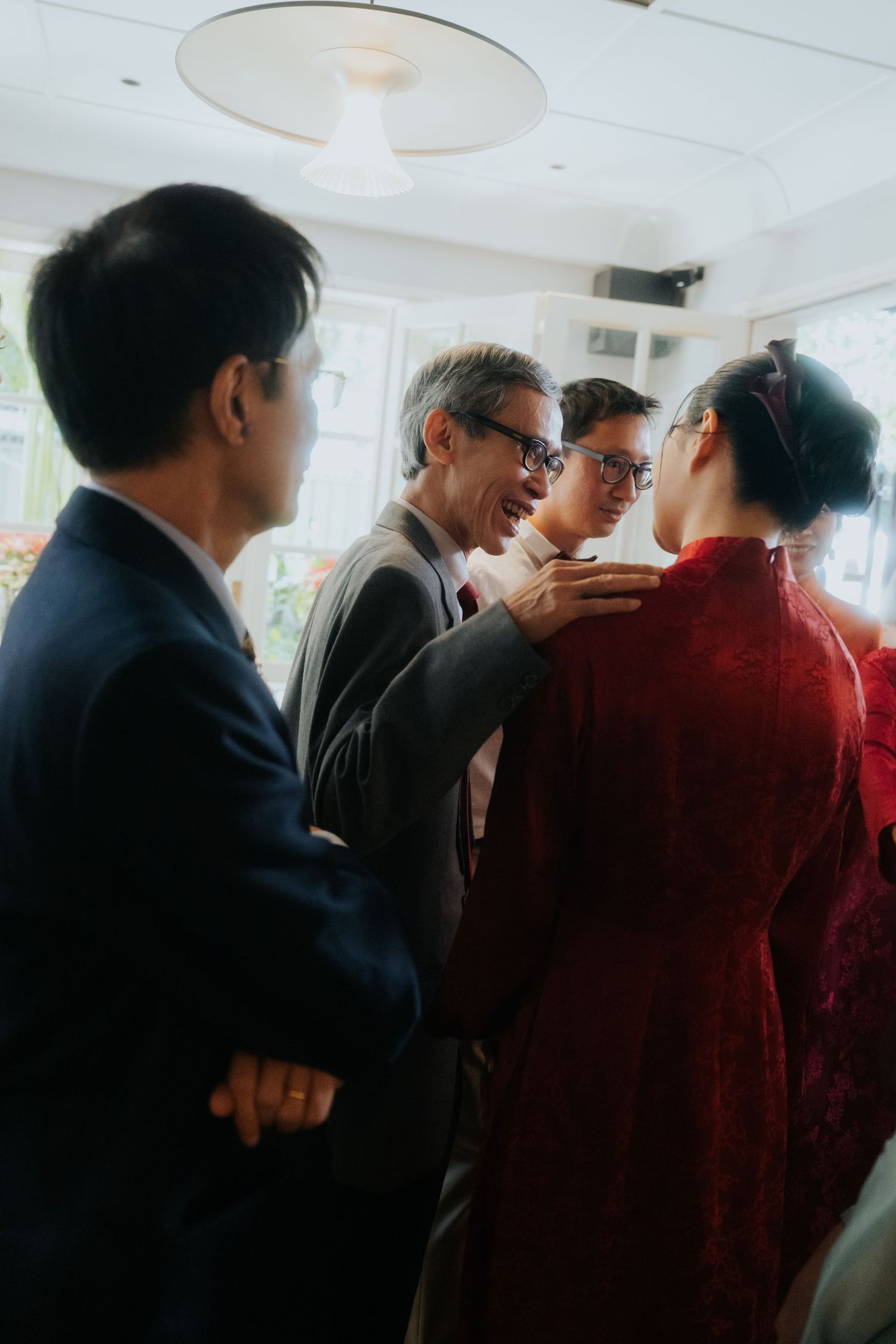 Guests in formal attire interact warmly at an Asian wedding ceremony, with a woman in a red traditional dress.