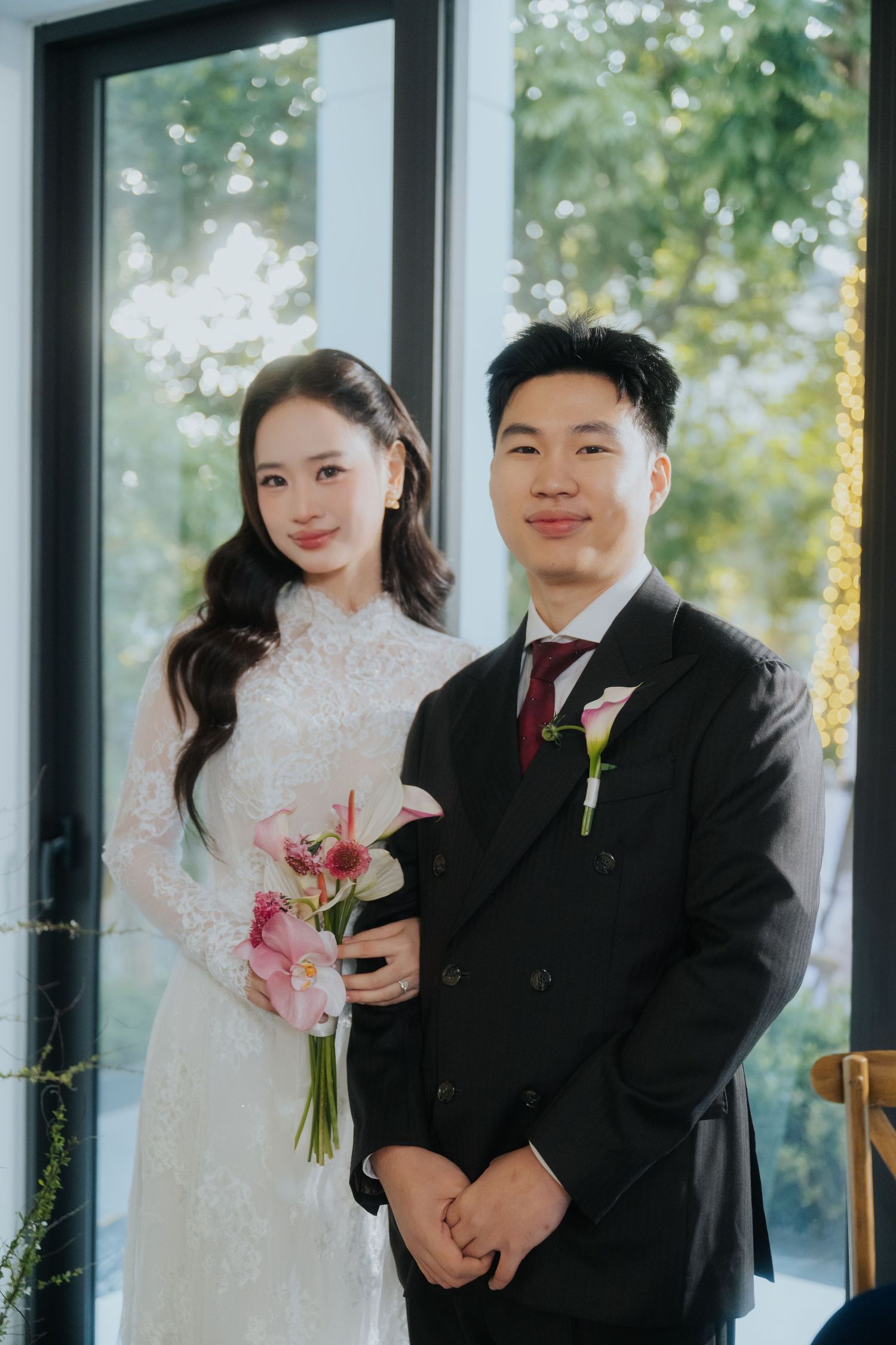 A bride in a white lace gown holding pink flowers stands beside a groom in a black suit with a boutonniere.