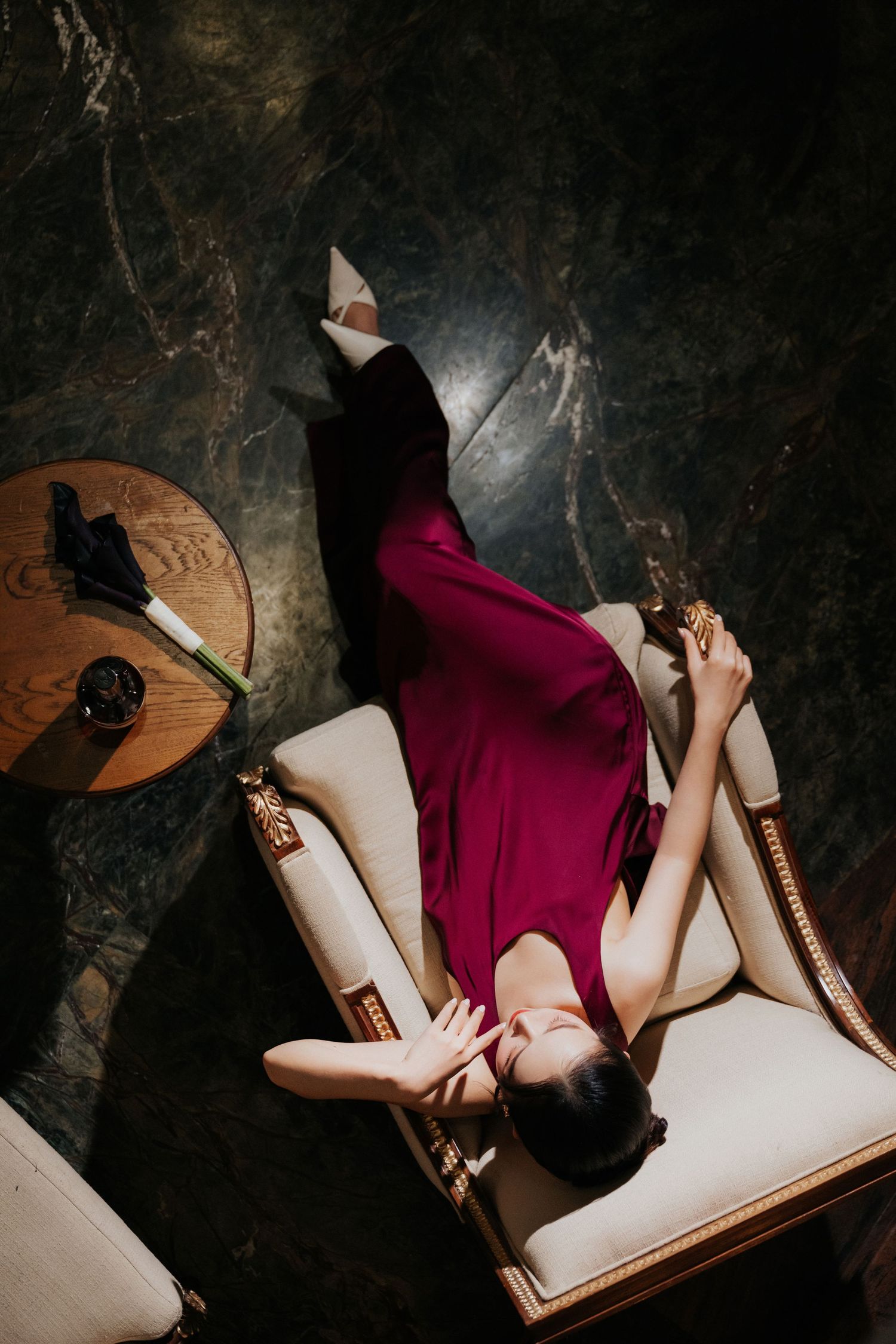 Woman in burgundy dress reclines gracefully on ornate white chair beside wooden table in dark elegant room.
