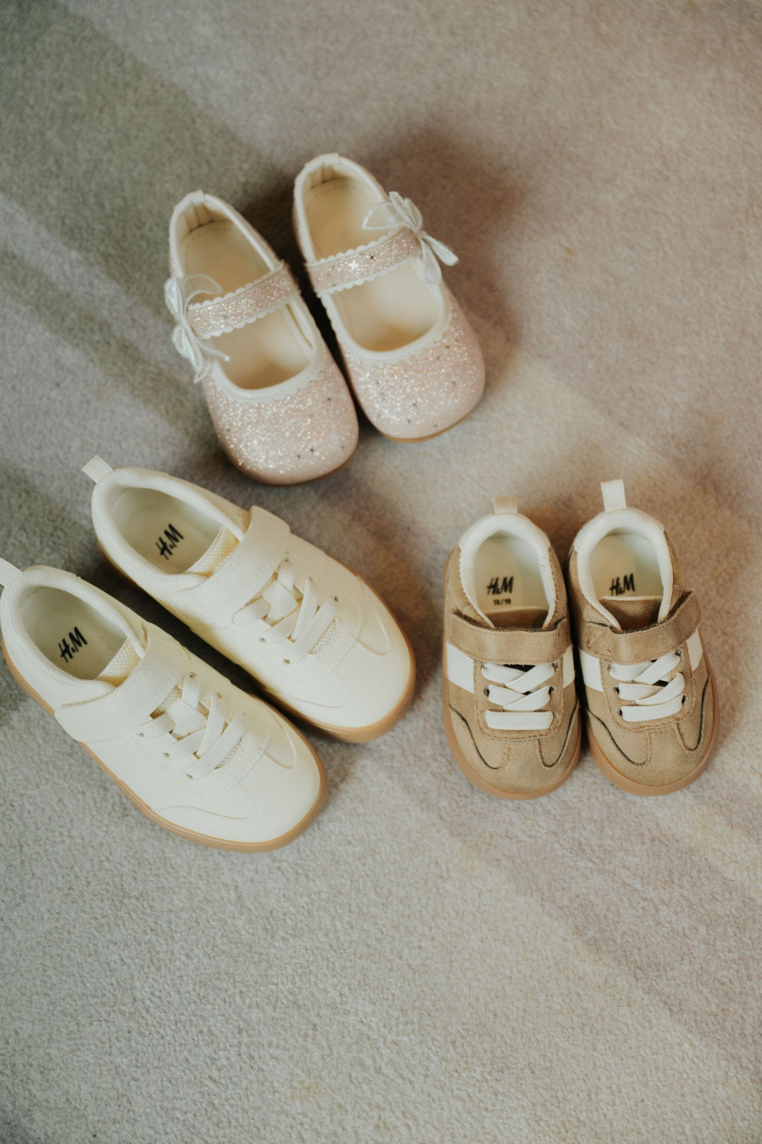 Three pairs of H&M kids shoes on carpet: white sneakers, tan suede sneakers, and pink glitter Mary Jane flats.