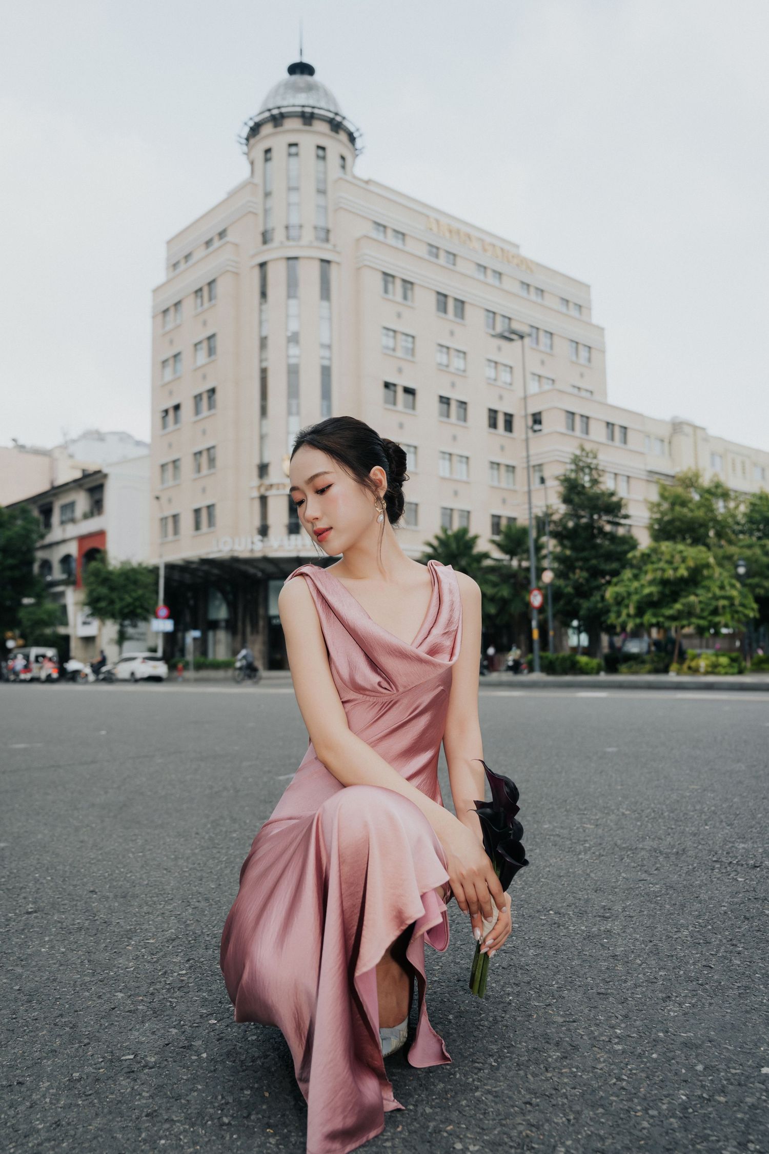Young Asian woman in pink satin slip dress sits on urban street holding flowers, with classic building backdrop.