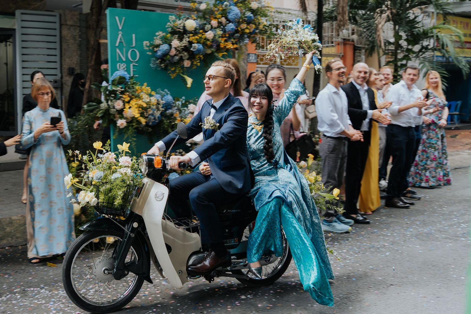 Joyful bride and groom riding a vintage moped through cheering guests on a decorated Vietnamese street wedding.