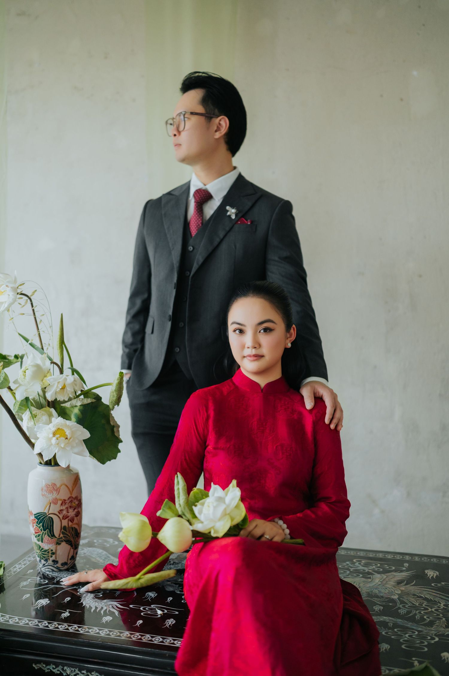 A couple in traditional Vietnamese attire, man in dark suit and woman in red ao dai holding white lotus flowers.