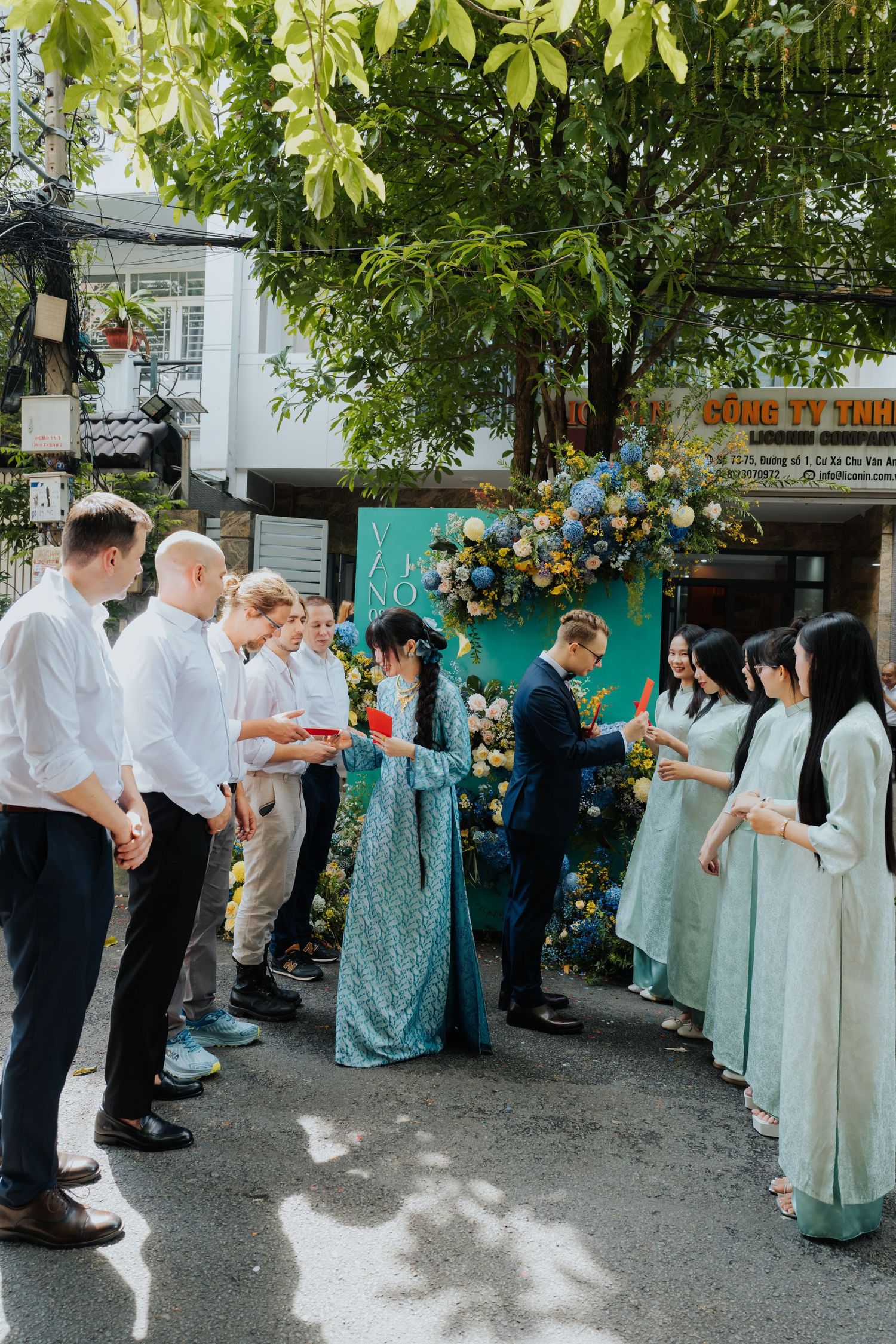 Vietnamese wedding ceremony outdoors with bride in blue ao dai, groom in suit, and guests exchanging gifts by floral arch.