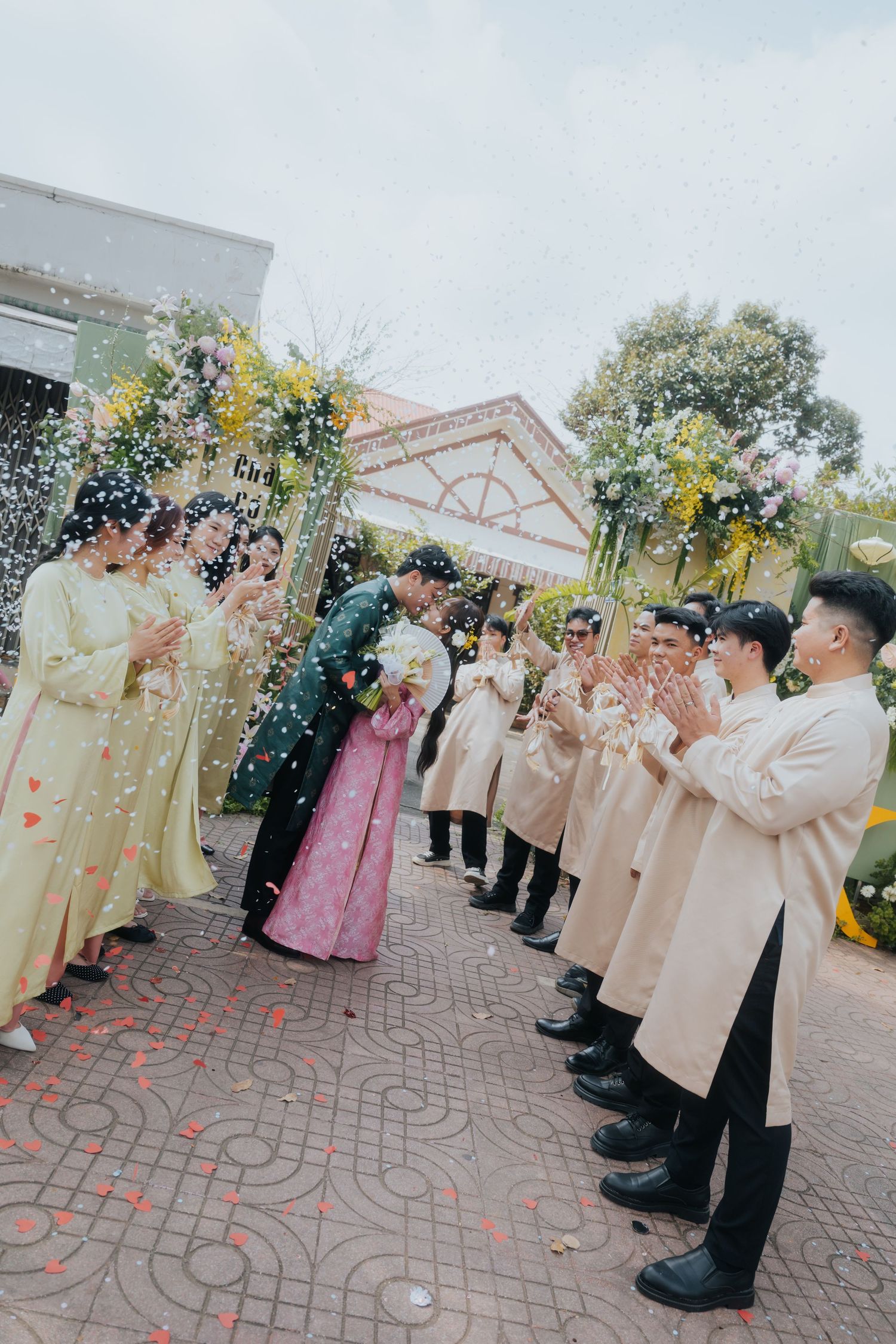 A couple kisses during a traditional Vietnamese wedding ceremony while guests in ao dai attire clap and confetti fills the air.