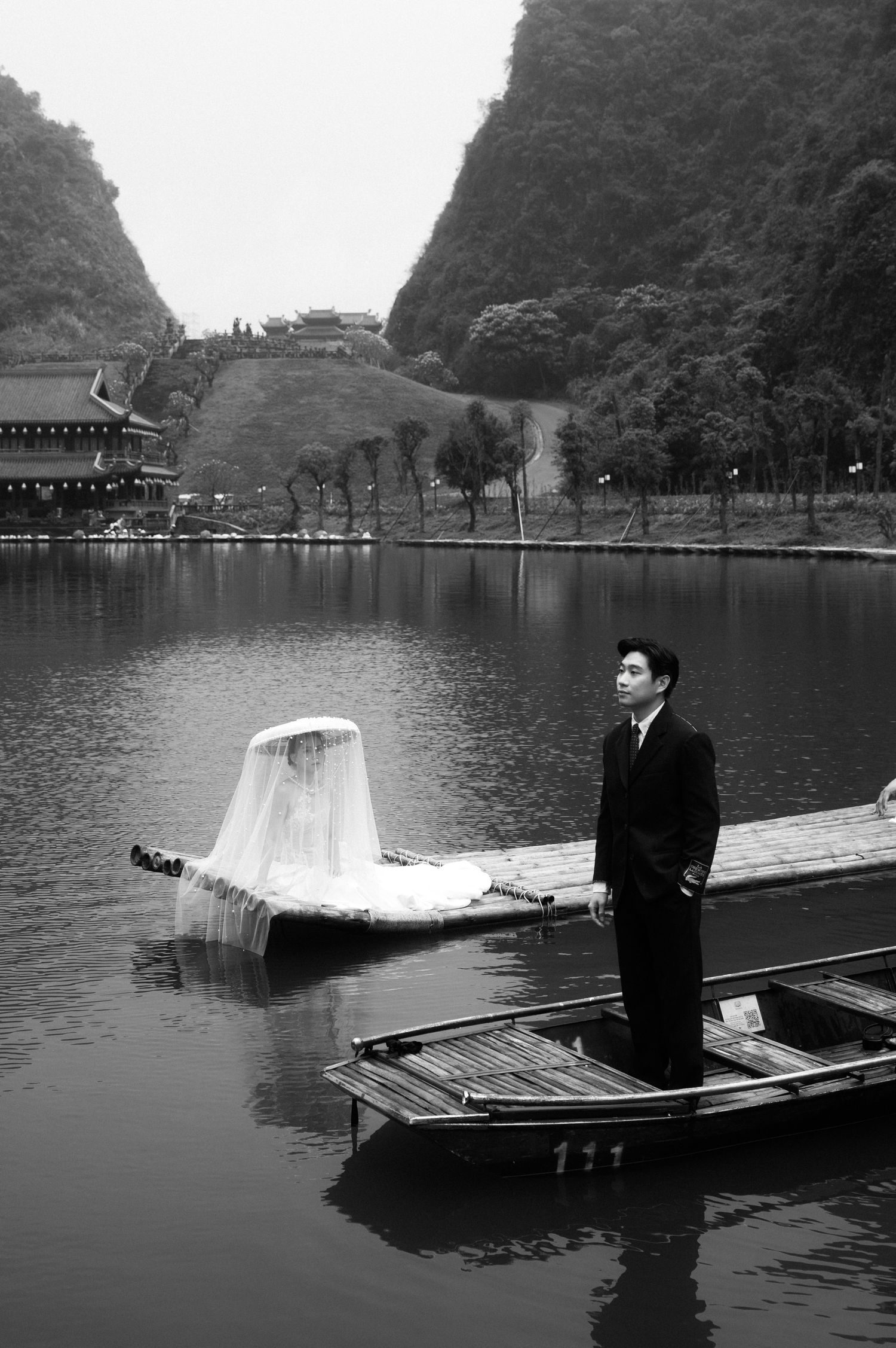 Black and white wedding photo of groom standing on wooden boat by scenic lake with karst mountains backdrop.