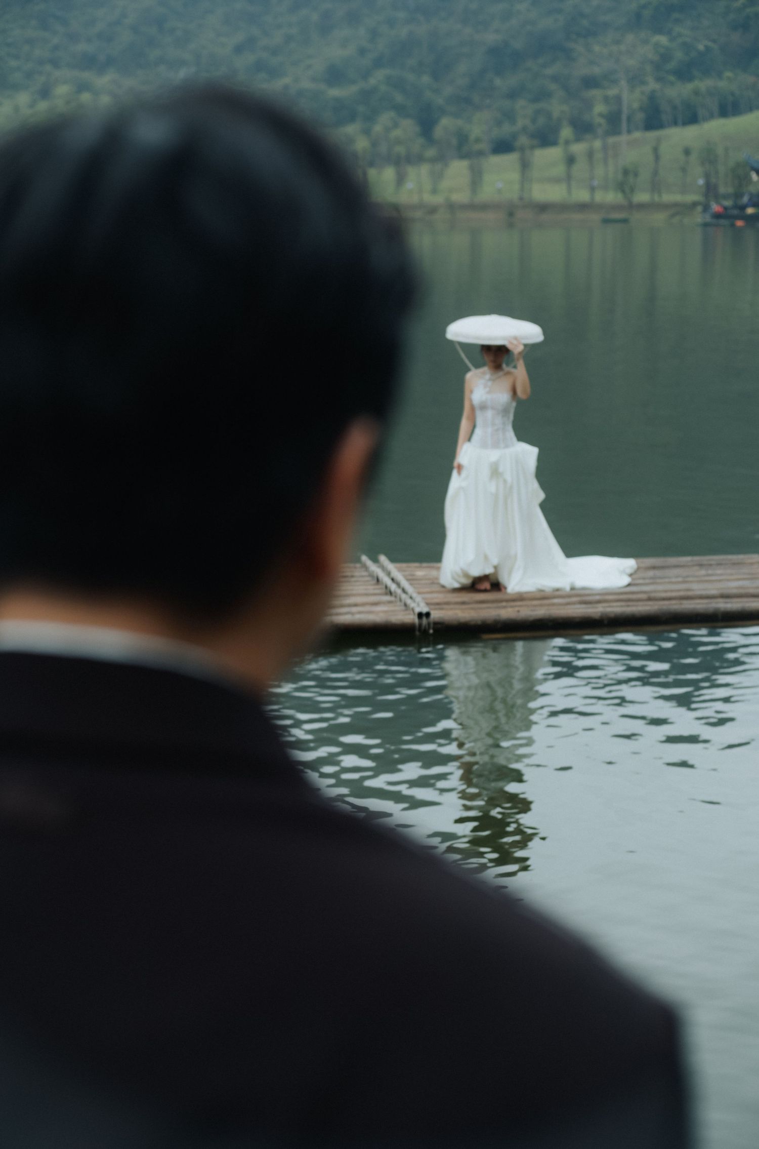 A groom watches his bride in a white dress holding an umbrella on a wooden dock over a serene lake.