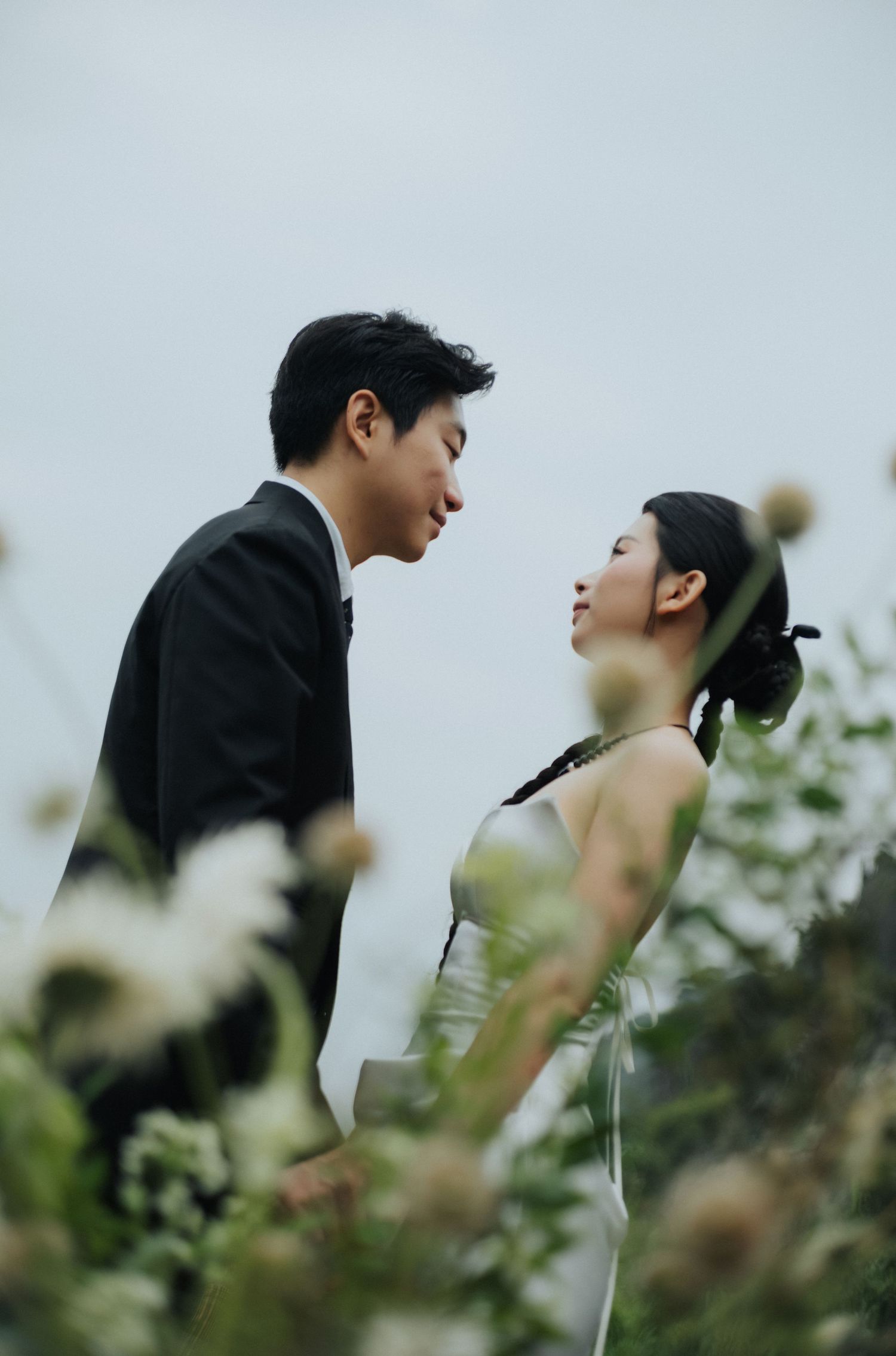 A couple in wedding attire shares a romantic moment surrounded by wildflowers against a soft gray sky.