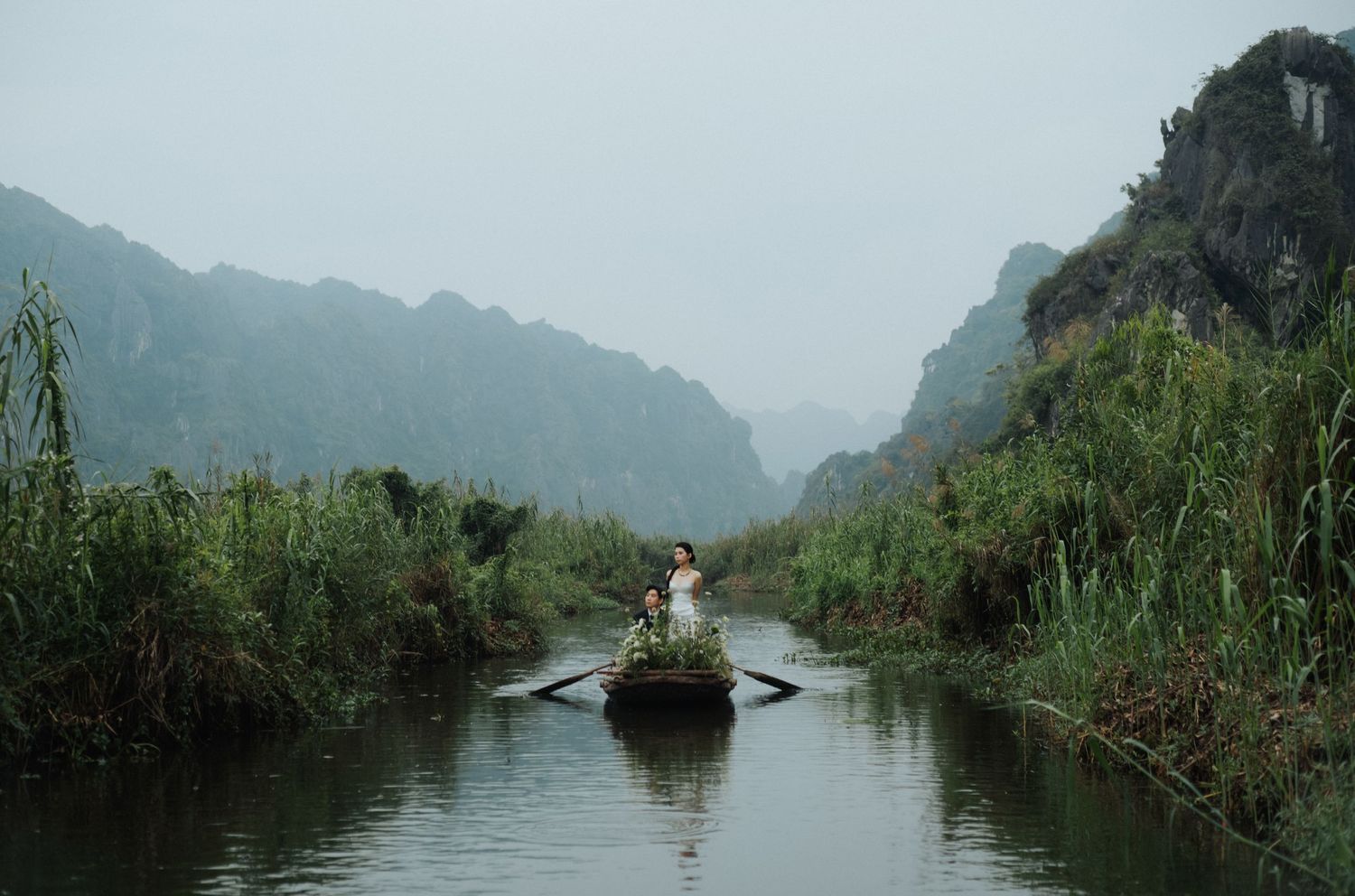 Person rowing a wooden boat through a misty river surrounded by lush greenery and karst mountains in Vietnam.