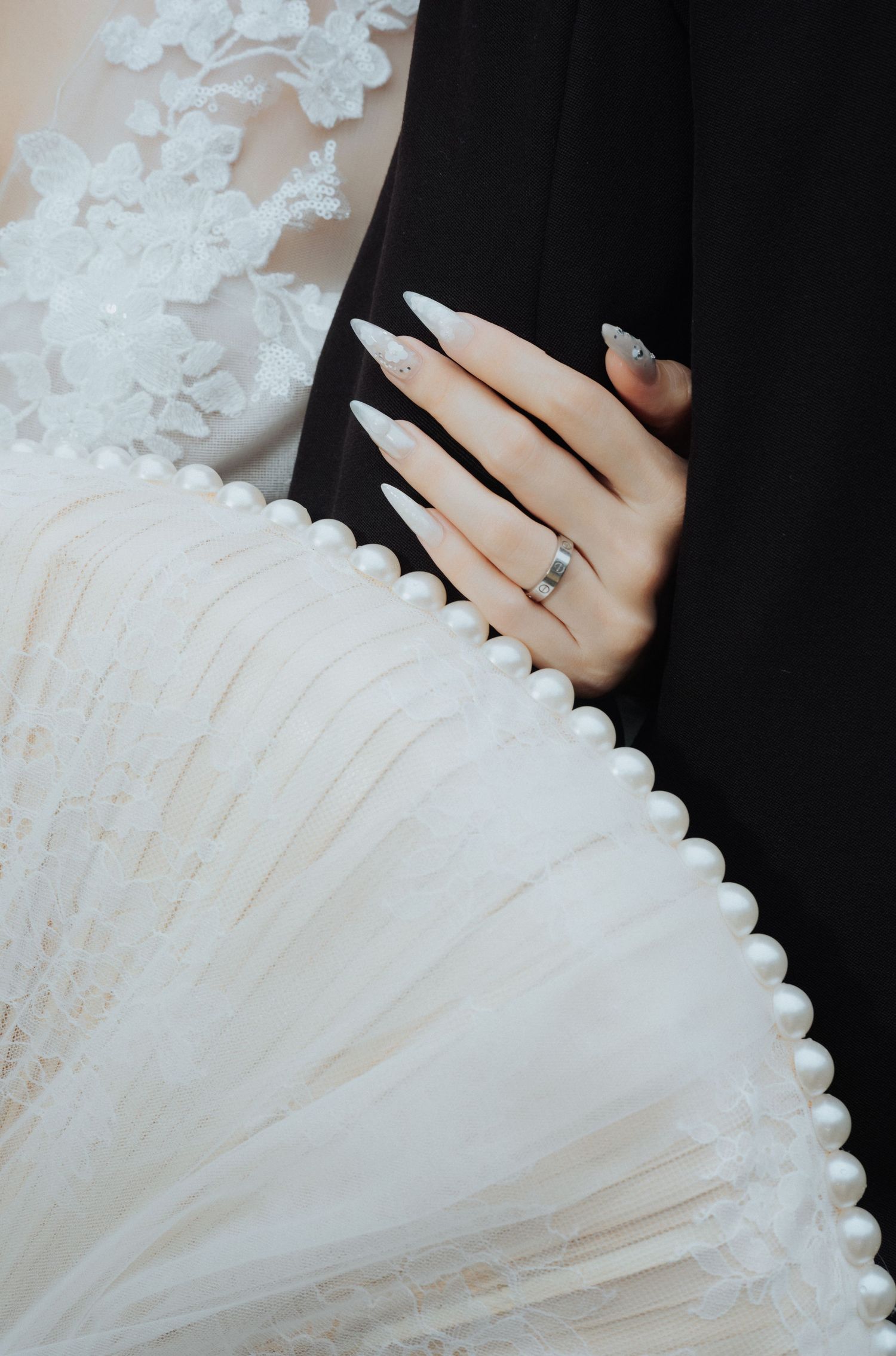 Bride's hand with long white stiletto nails and silver ring resting on groom's black suit beside pearl-trimmed wedding dress.