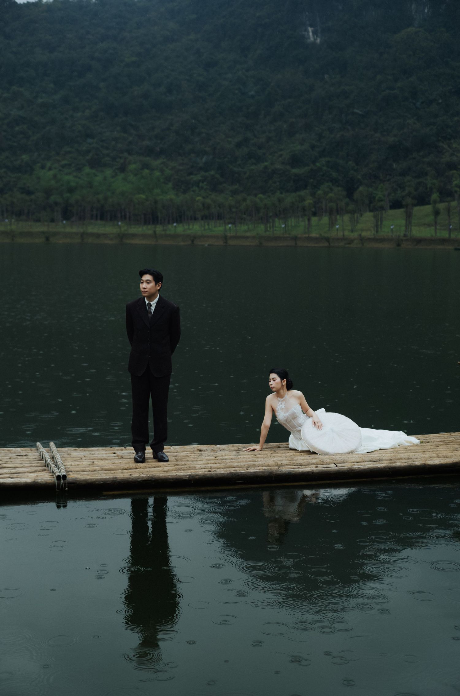 A bride in a white gown reclines on a dock while a groom in a black suit stands nearby over a serene lake.