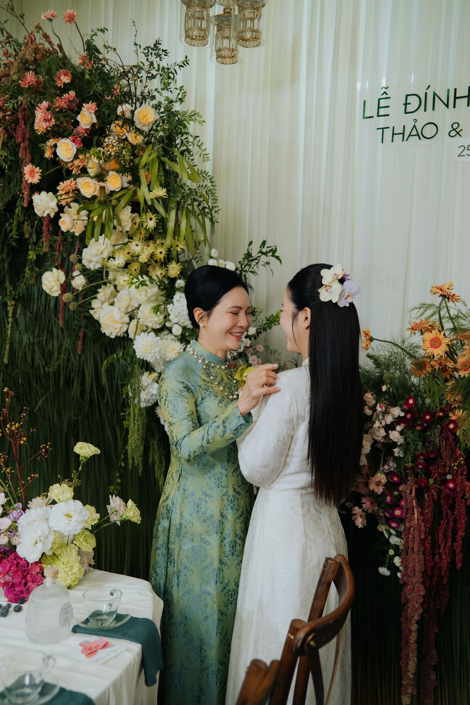 Two women in ao dai share a joyful moment at a Vietnamese wedding ceremony surrounded by lush floral arrangements.