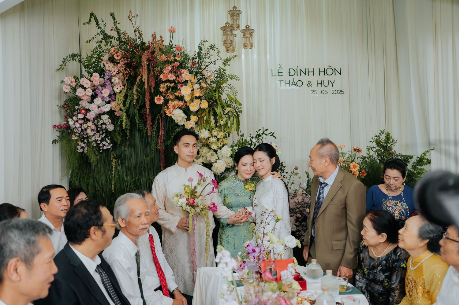 Vietnamese engagement ceremony with couple in traditional ao dai attire, surrounded by family at a floral-decorated reception venue.