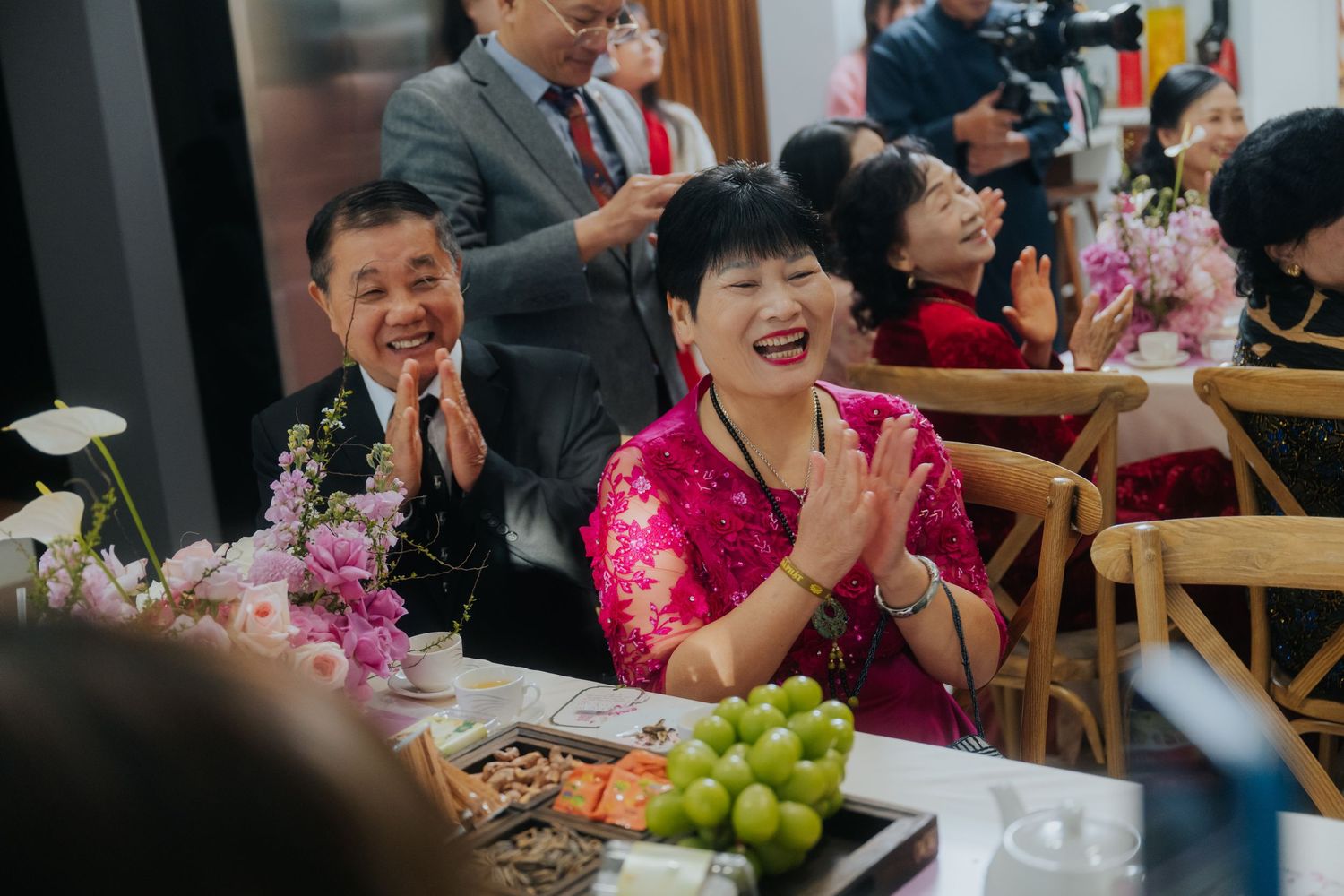 Joyful Asian guests clapping and laughing at a festive celebration with flowers and fruit on the table.