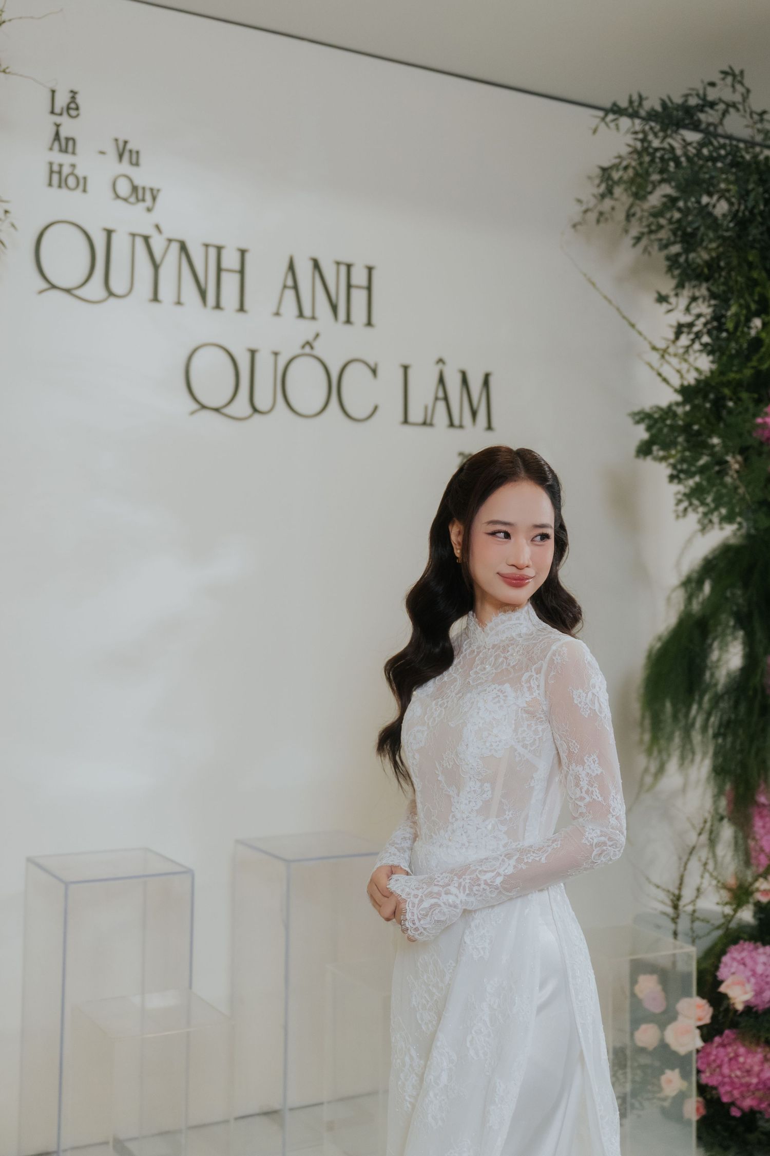 A bride in a white lace ao dai gown stands before a wedding backdrop displaying Quynh Anh and Quoc Lam.
