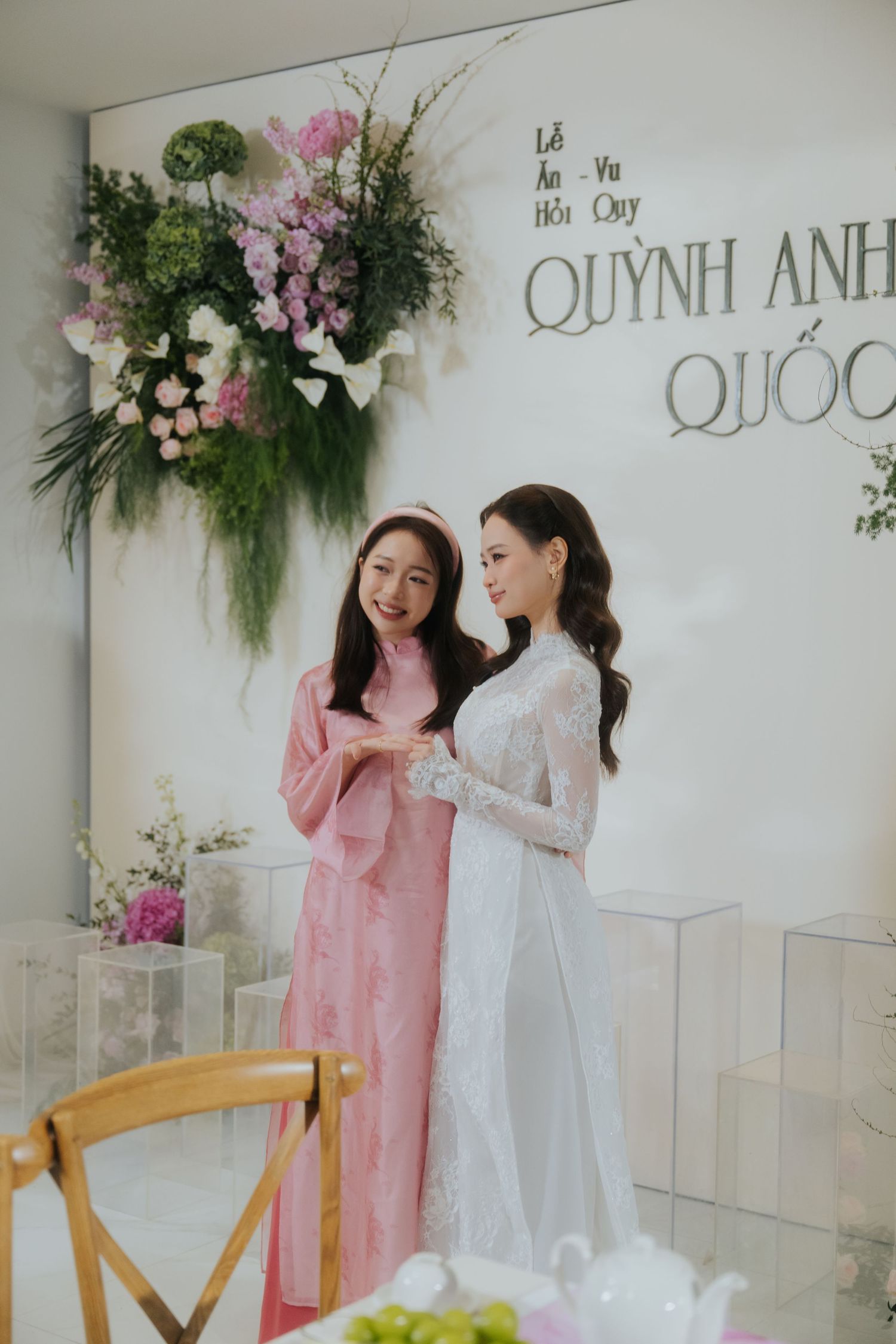 Two women in elegant ao dai dresses pose at a Vietnamese engagement ceremony decorated with floral arrangements.