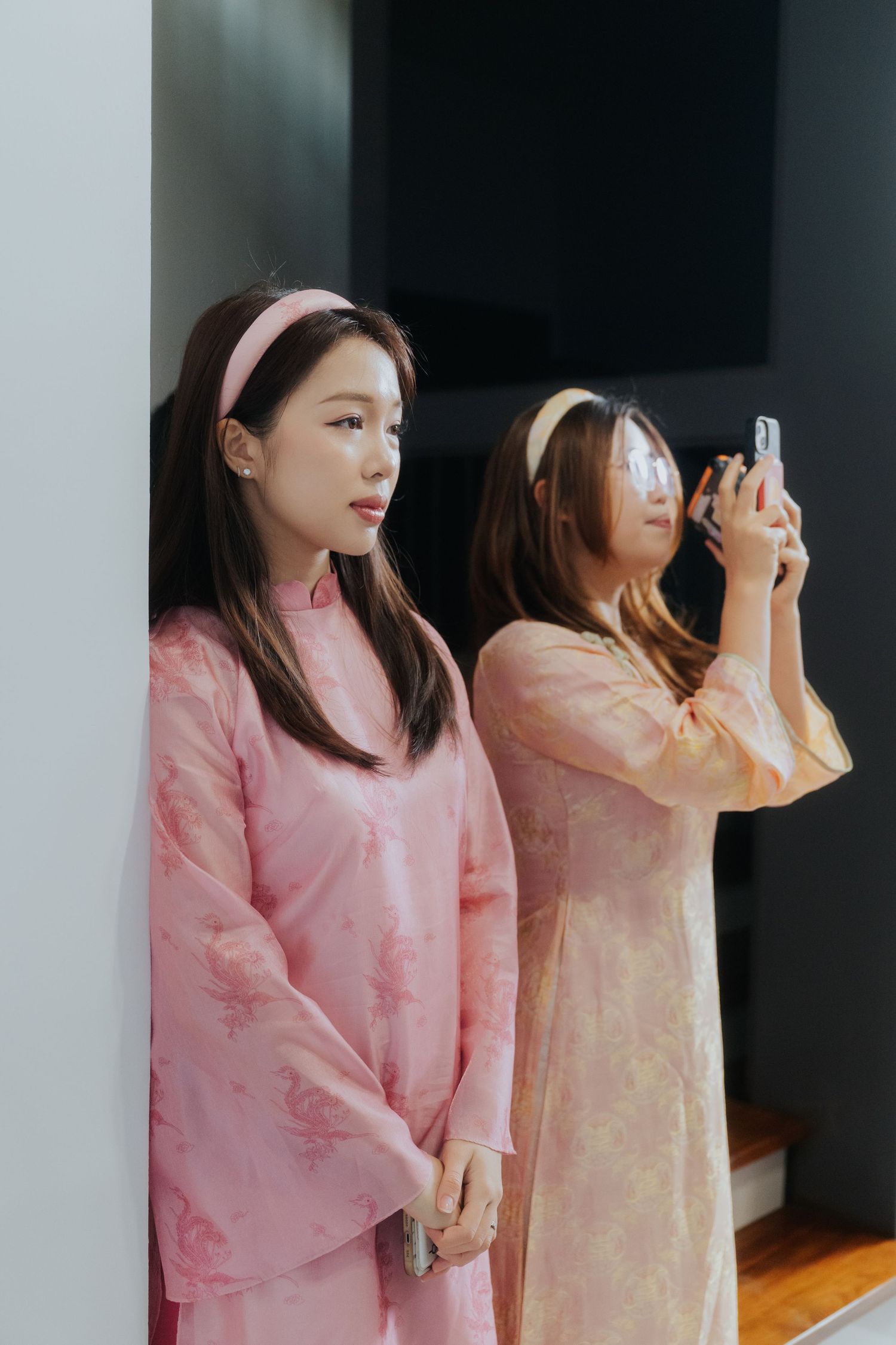 Two young women in matching pink floral ao dai dresses, one taking a photo with smartphone.