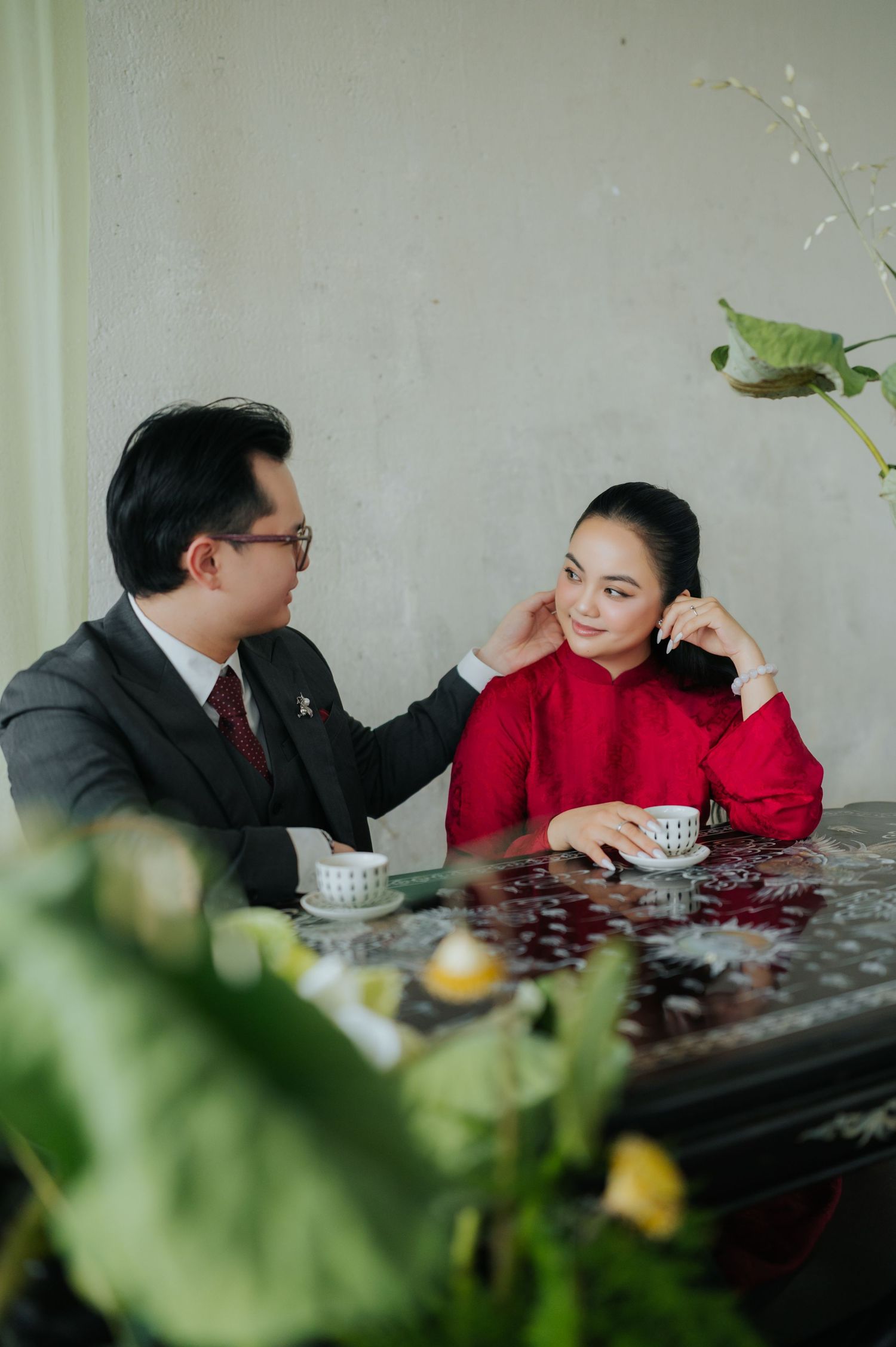 A couple in elegant Vietnamese traditional outfits share a tender moment at a decorative table with tea cups.