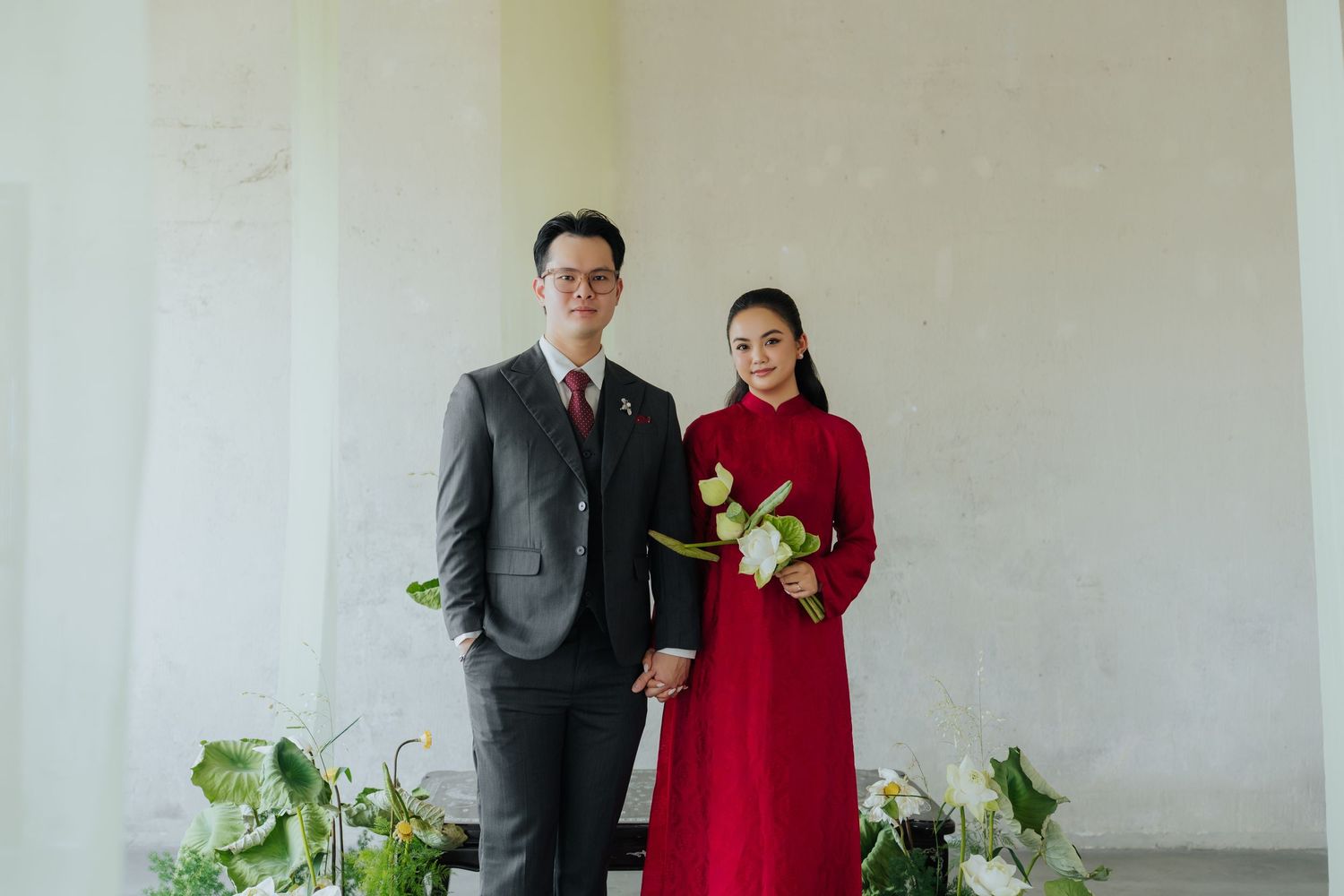 Couple in formal attire, man in gray suit, woman in red ao dai holding white flowers, posing indoors.