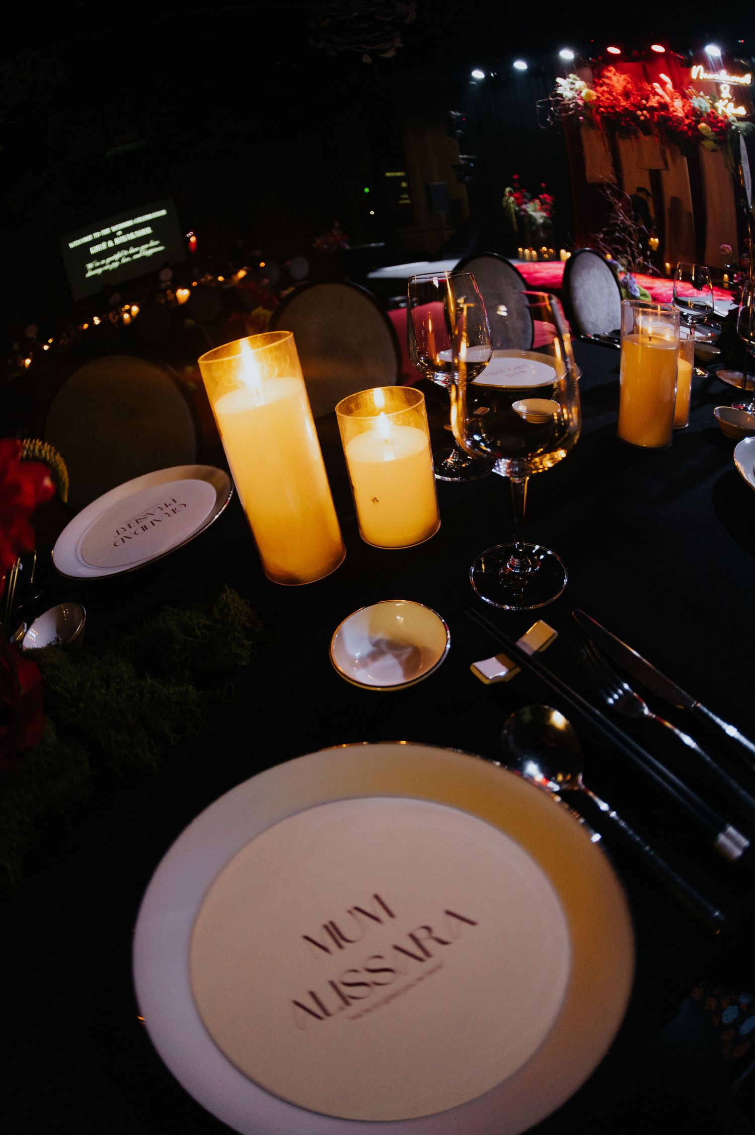 Elegant candlelit dinner table setting with glowing pillar candles, wine glasses, and a personalized place card plate.