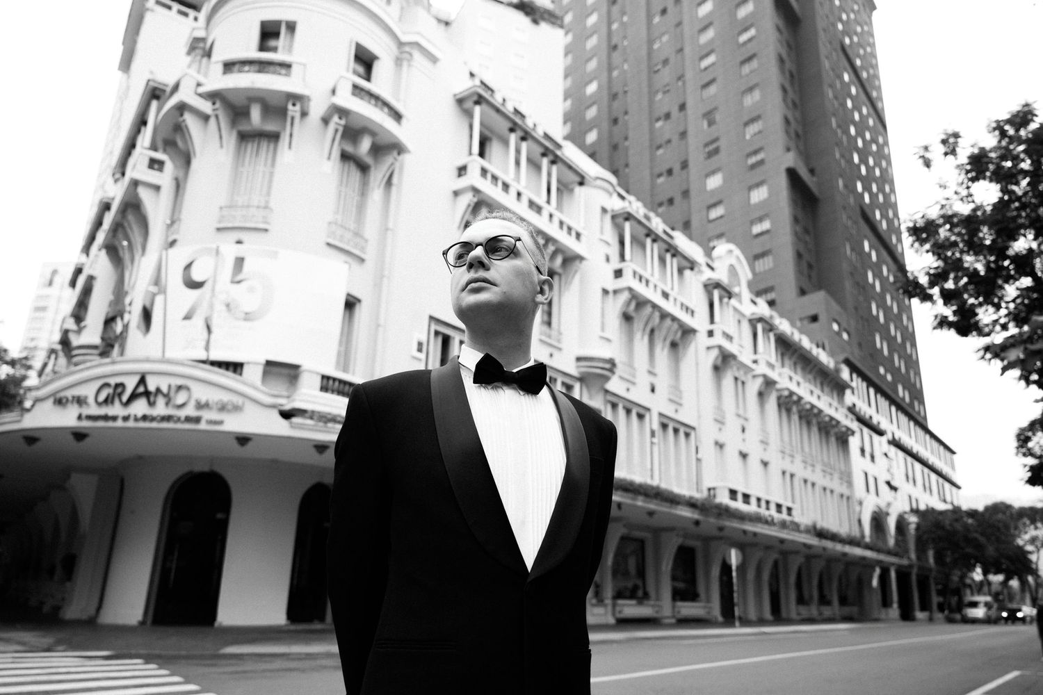 Man in tuxedo poses on street in front of Hotel Grand building in black and white photo.