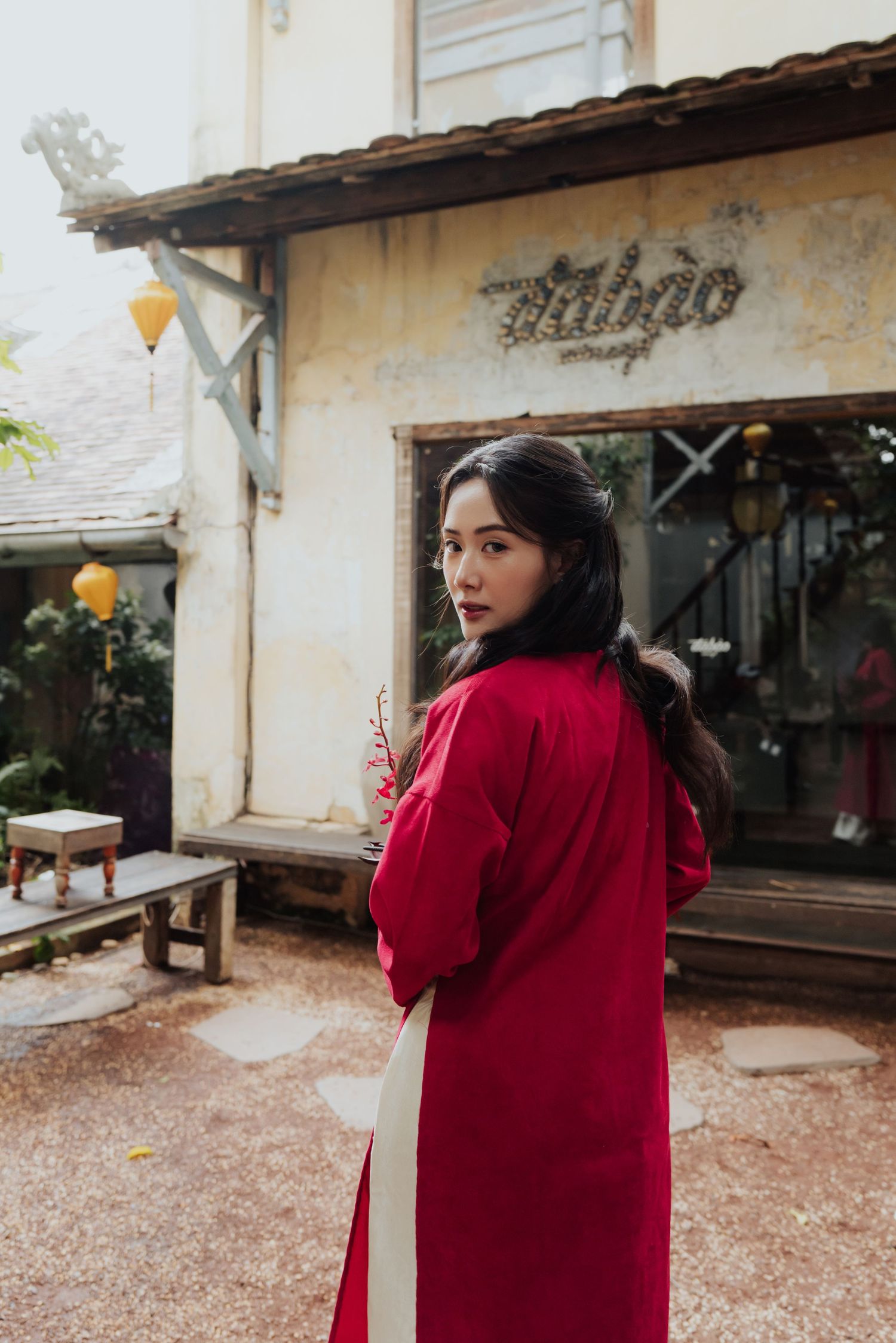 Young woman in red ao dai traditional Vietnamese dress poses outside a rustic cafe with yellow lanterns.