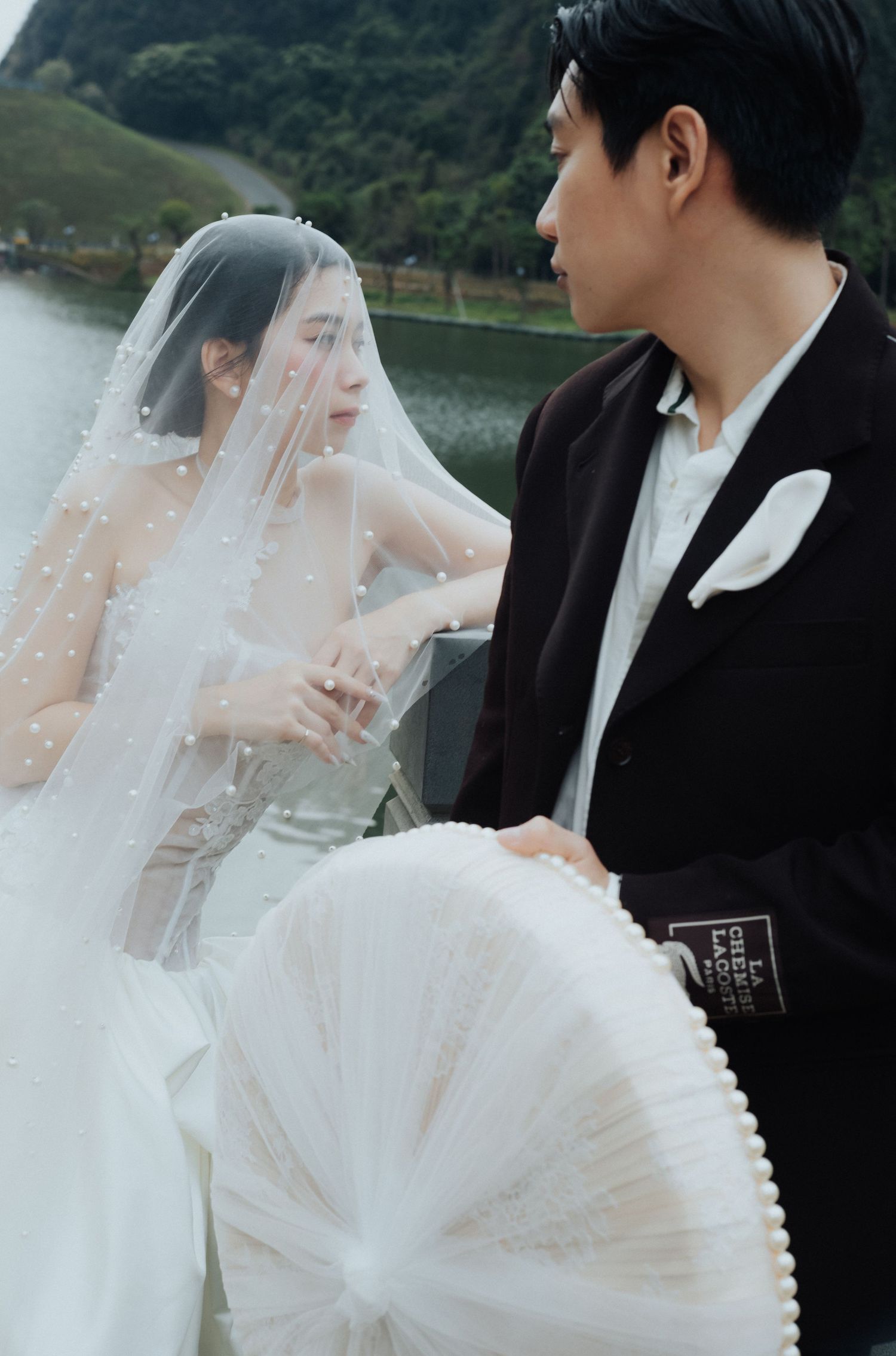 A bride in a pearl-adorned veil and groom in a black tuxedo share an intimate moment by a serene lake.