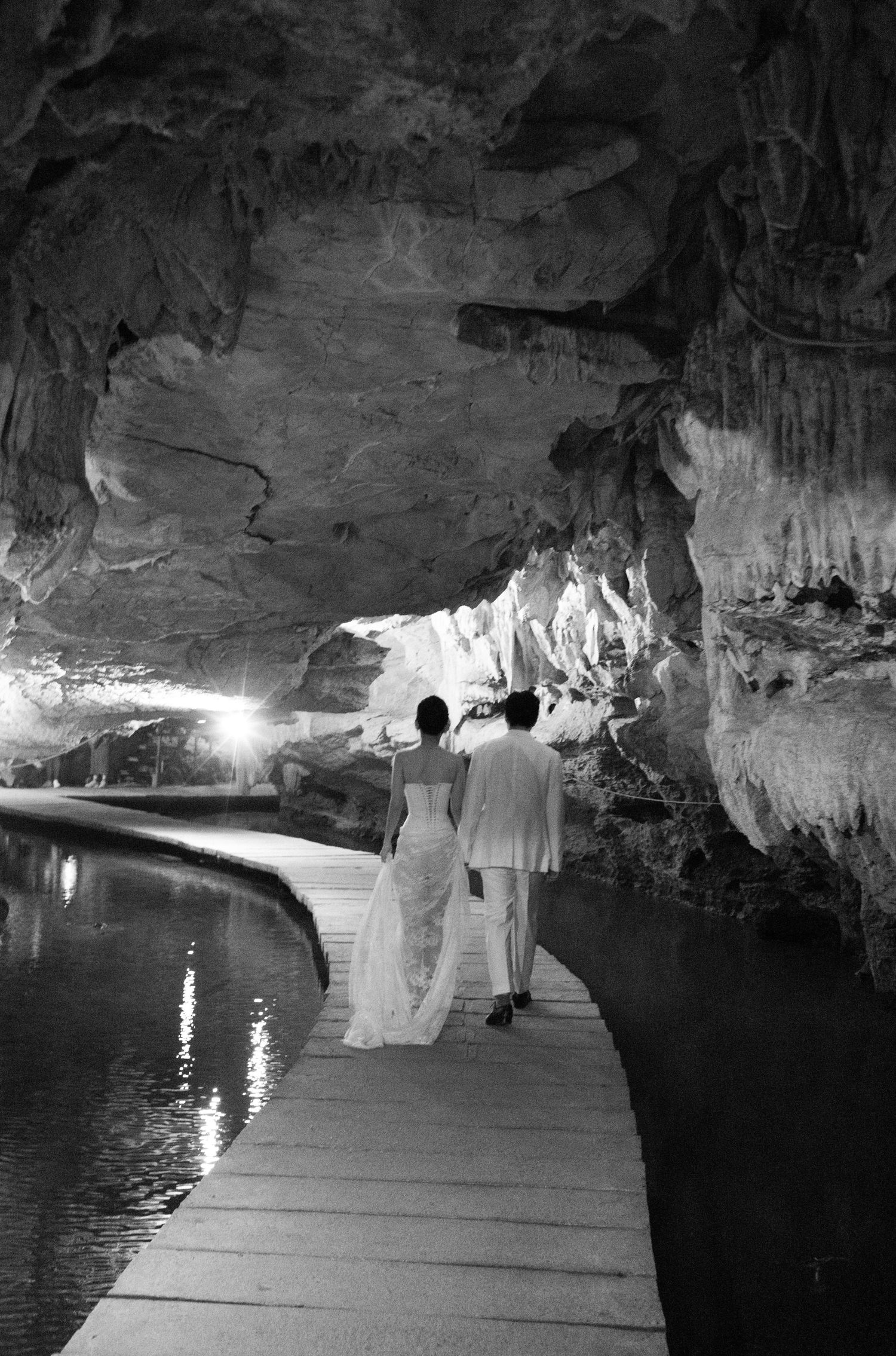 Bride and groom walk hand-in-hand along a wooden boardwalk inside a stunning cave with a reflective pool.