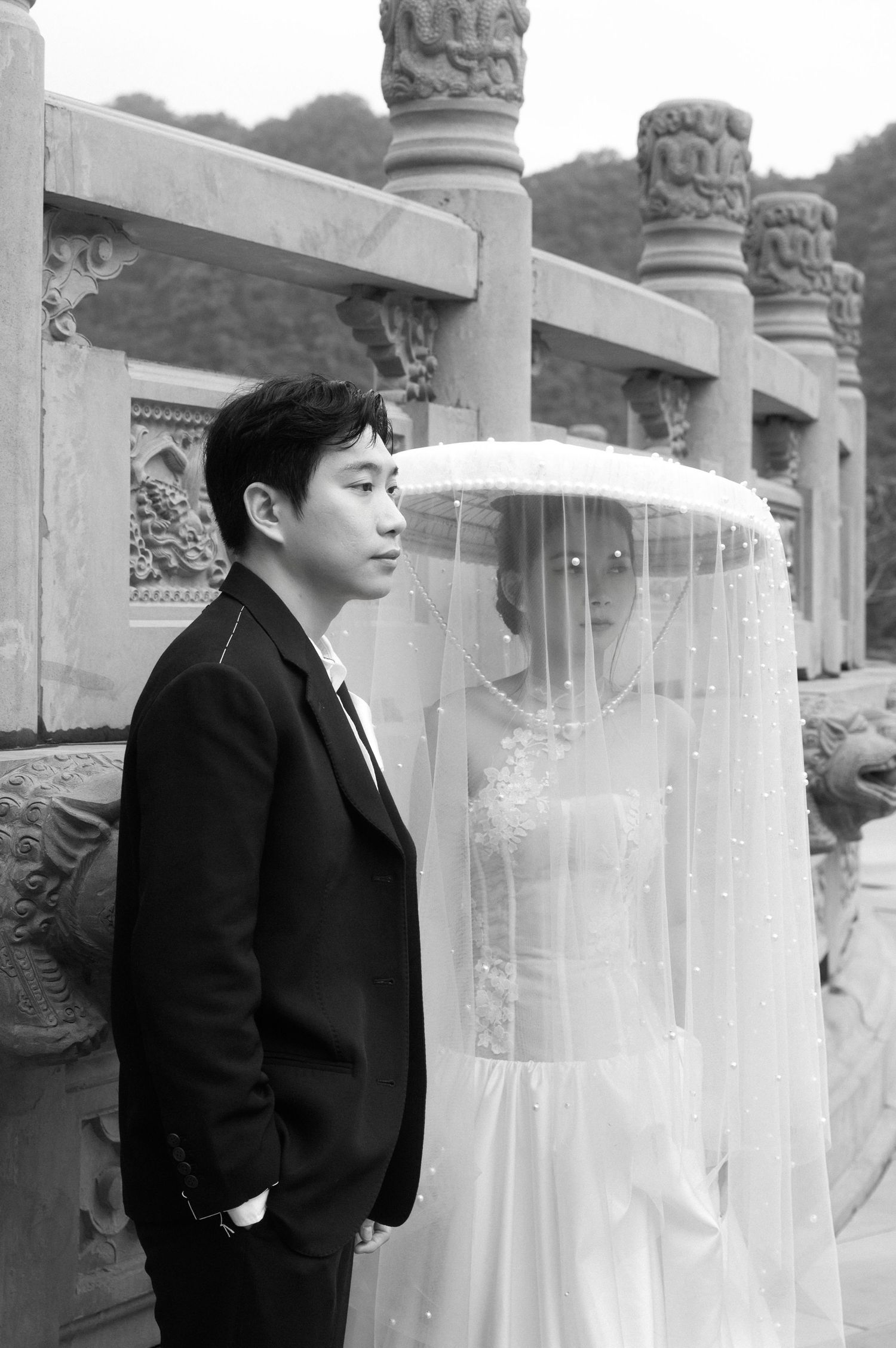 Black and white wedding photo of an Asian couple posing at ornate stone balustrade, bride in veil and groom in suit.