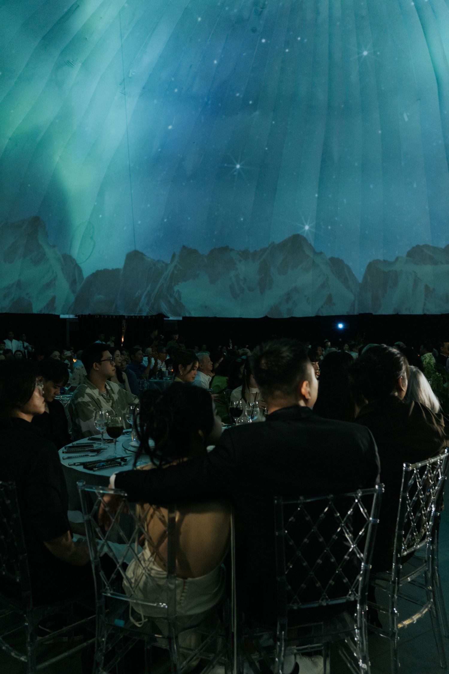 Guests dining under a stunning aurora borealis projection at an elegant gala event with acrylic chairs.