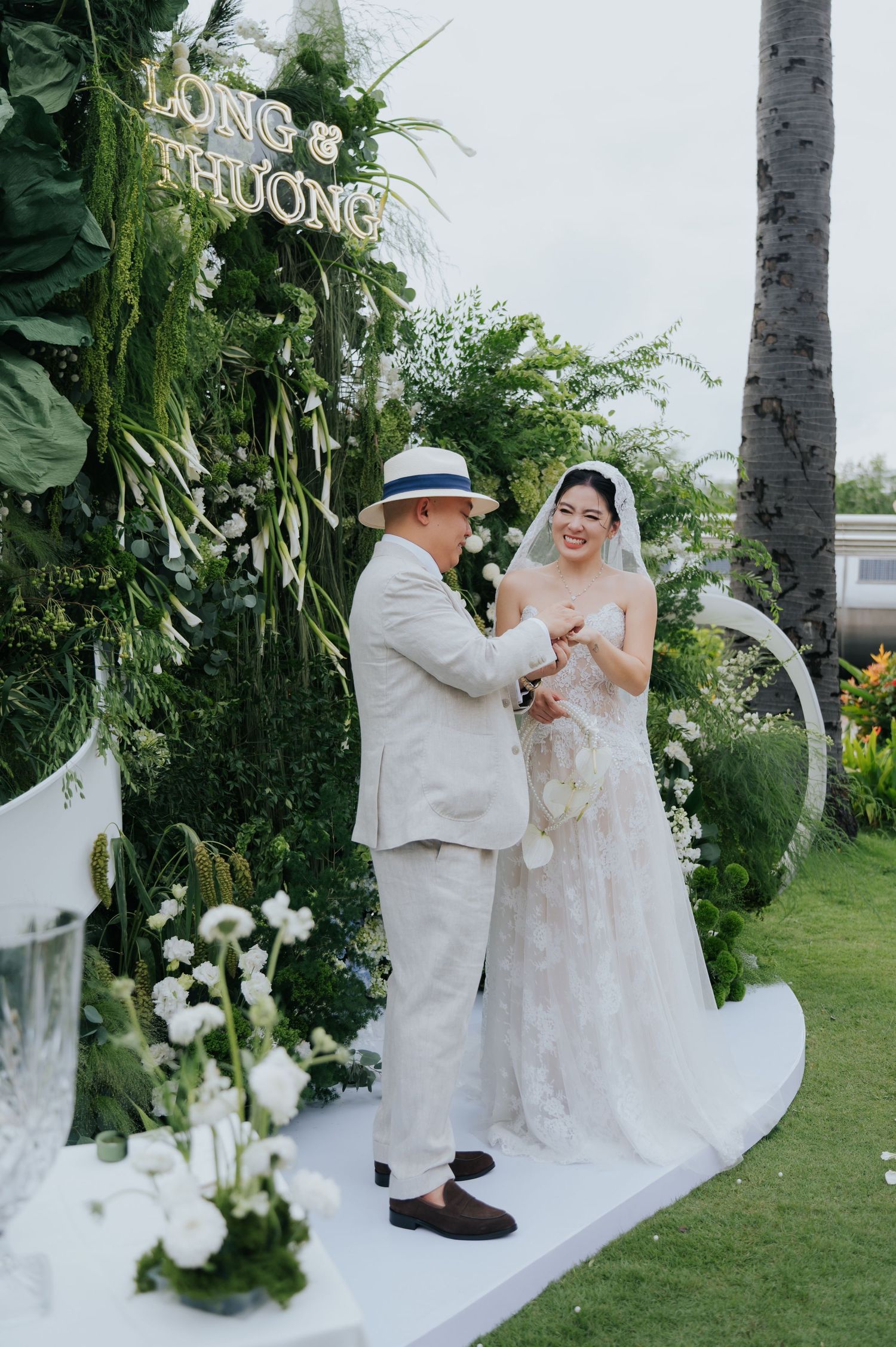 Bride and groom exchange vows at outdoor garden wedding ceremony with lush green floral backdrop and white decorations.