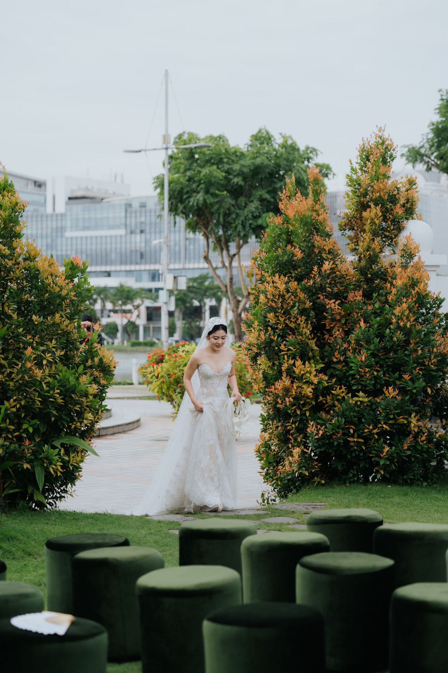 Bride in white lace gown and veil walks through lush garden with green ottomans in foreground at outdoor wedding venue.