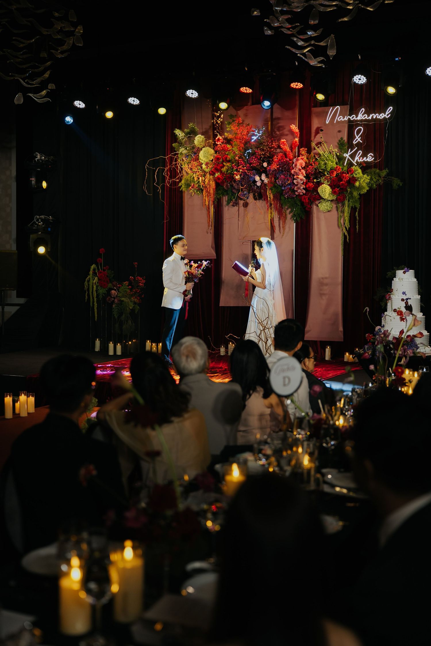 Elegant wedding reception with bride and groom at floral backdrop, candlelit tables, and neon name sign in dark venue.