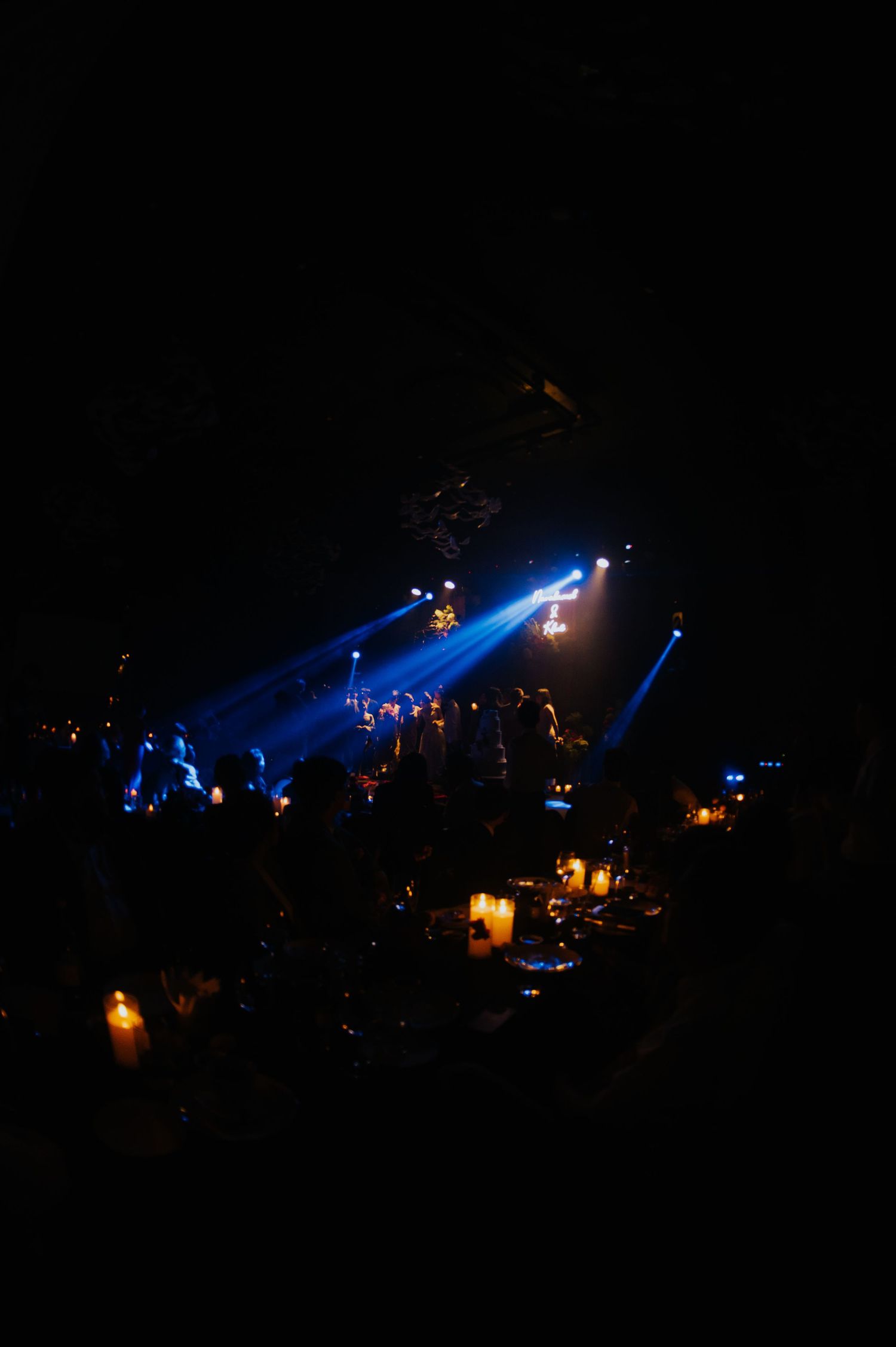 Dark venue with blue stage lights illuminating a live performance while guests dine by candlelight.