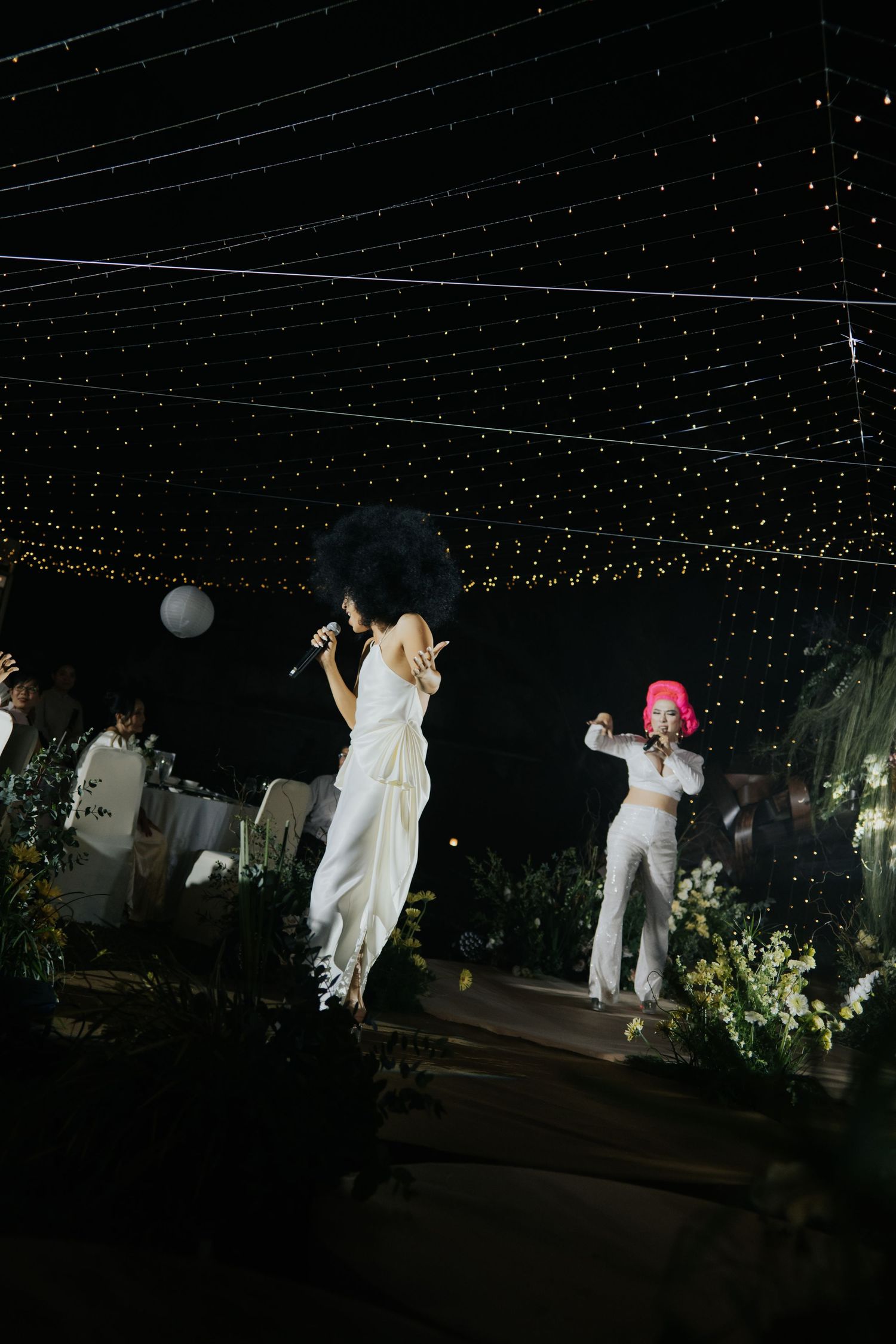 Two performers in white outfits sing on an outdoor stage adorned with fairy lights at a night event.