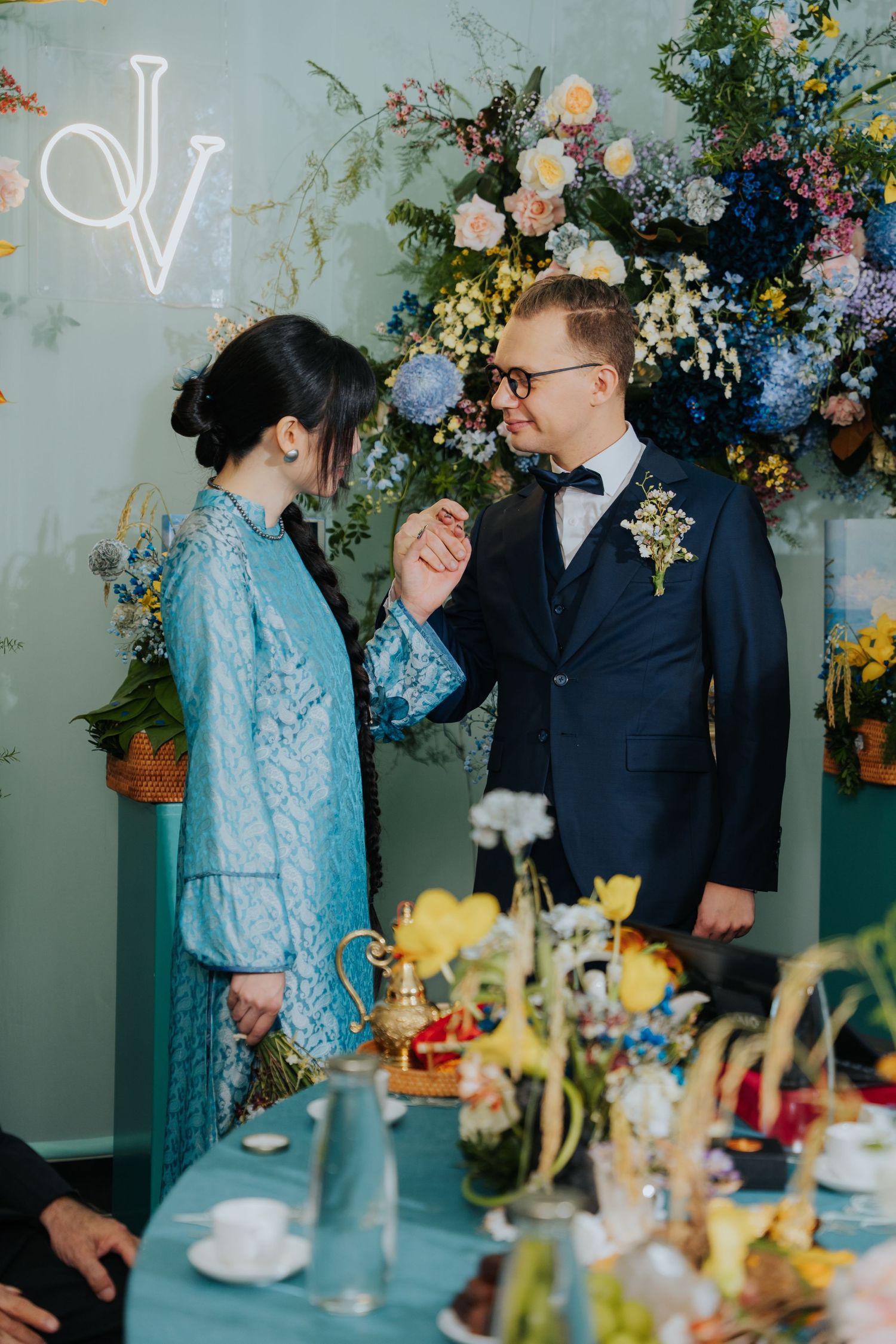 Bride in blue lace dress and groom in navy suit hold hands at floral-decorated wedding reception table.