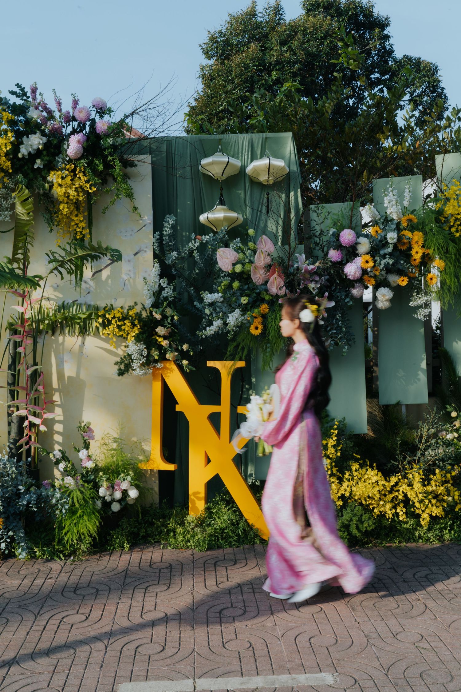 Woman in pink ao dai walks past floral wedding backdrop with yellow NK monogram letters and colorful blooms.