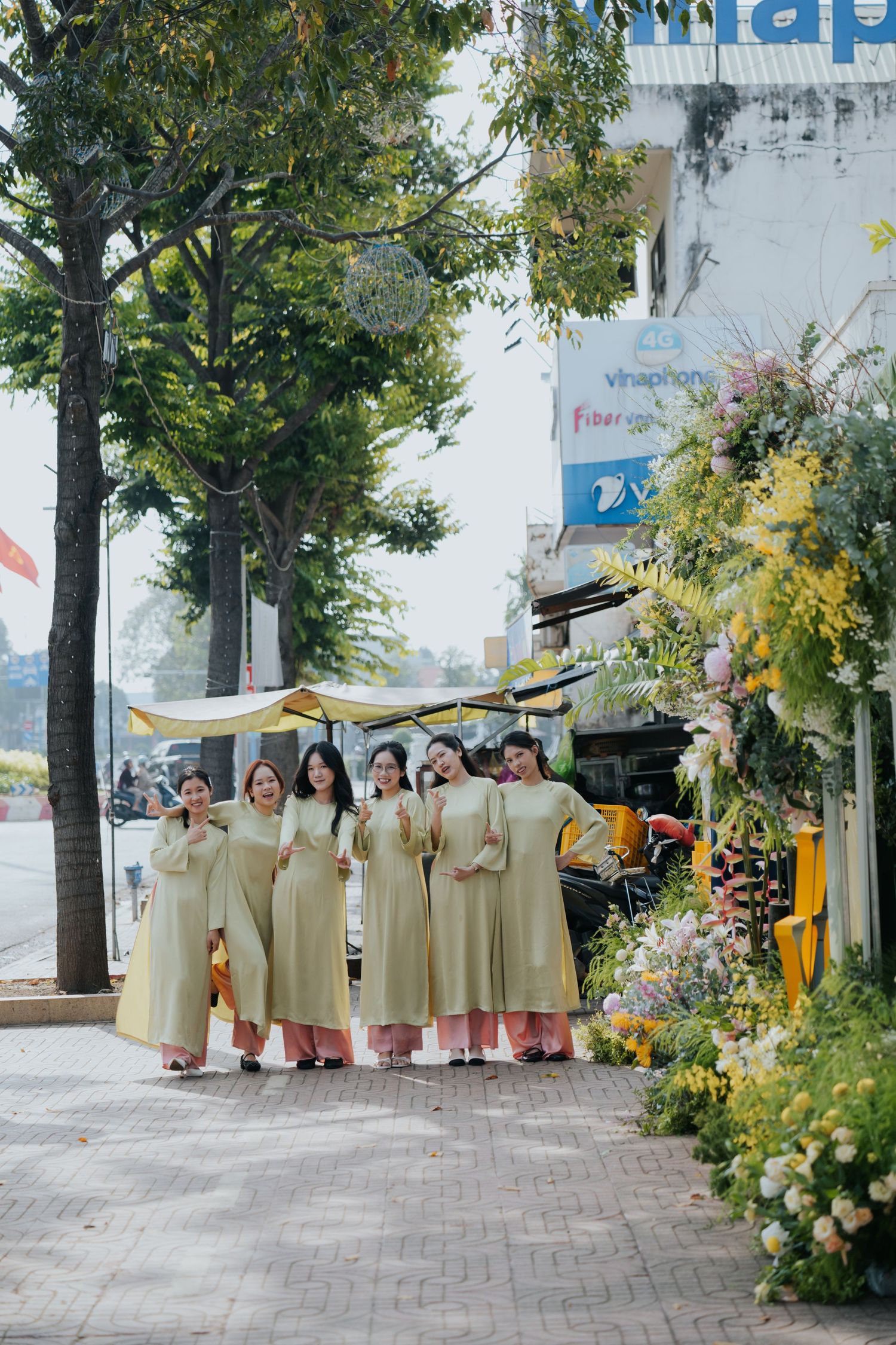 Group of women in matching yellow ao dai dresses posing on a Vietnamese street lined with flowers and trees.