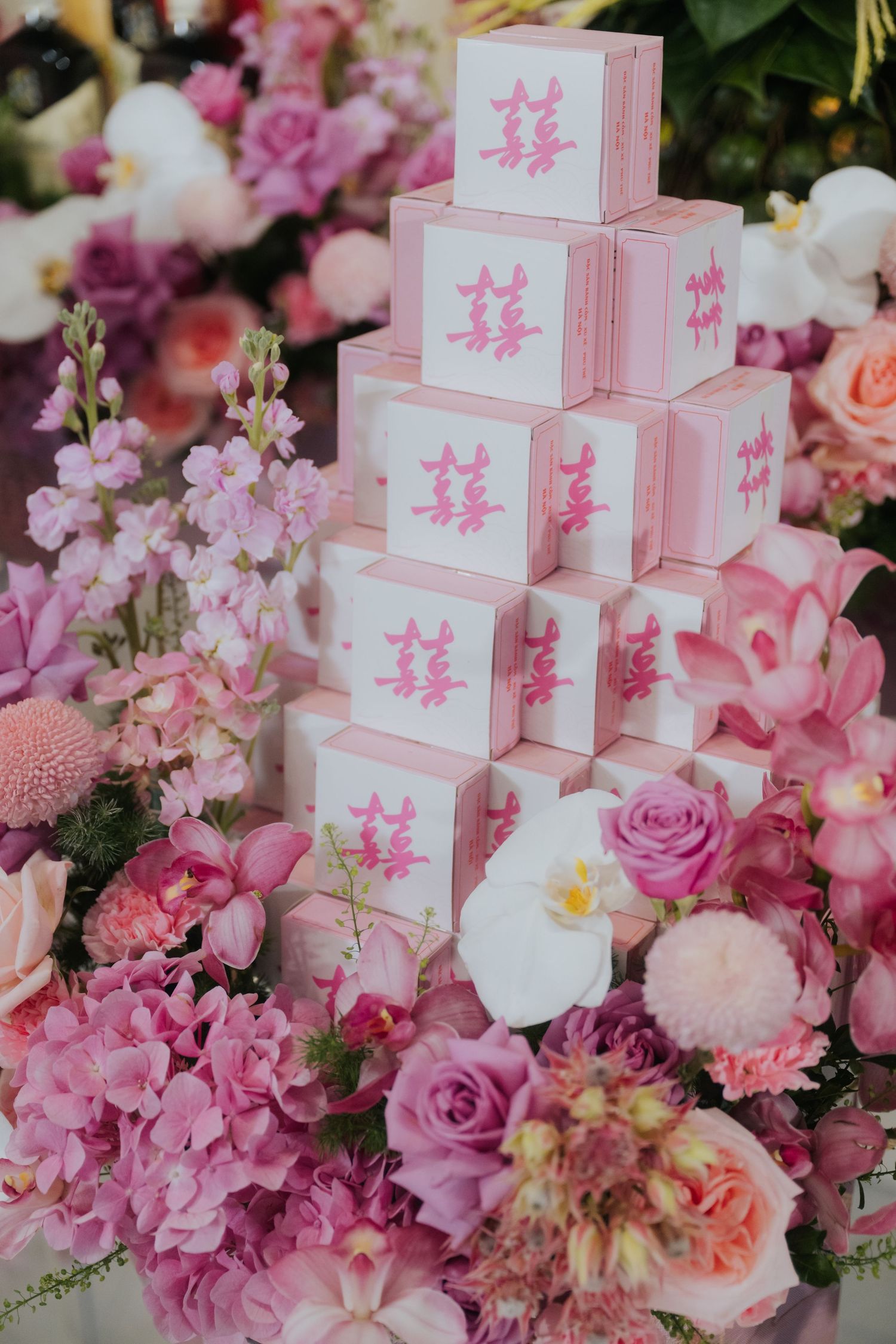 Pink and white tiered boxes with Chinese double happiness symbols surrounded by lush pink floral arrangements at a wedding.