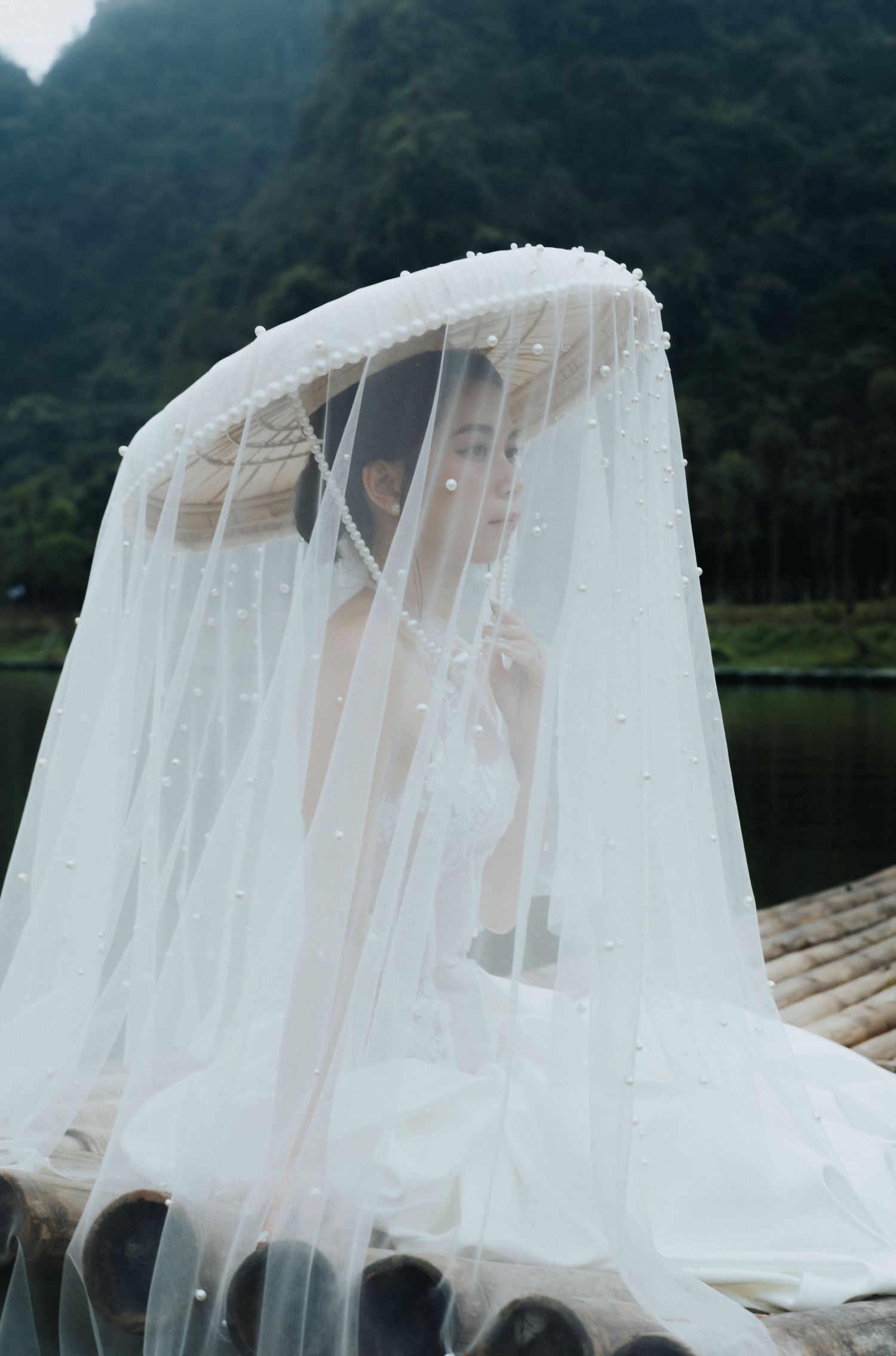 Bride in white wedding dress and pearl-adorned veil over conical hat, seated on bamboo raft with misty mountains backdrop.