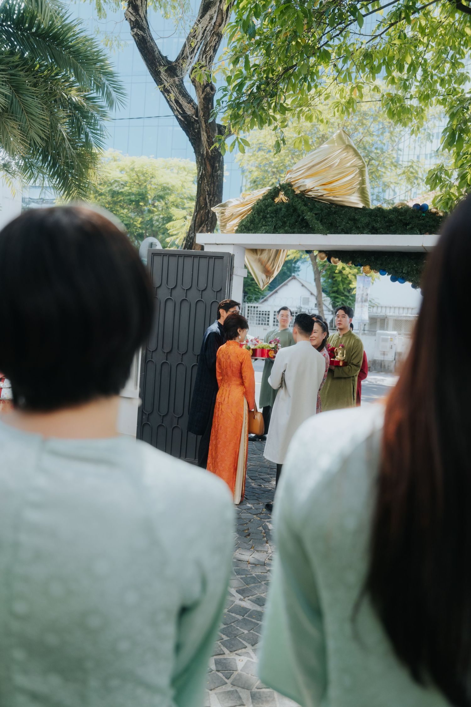 Guests in ao dai gather outdoors for a traditional Vietnamese wedding ceremony near a gate surrounded by trees.