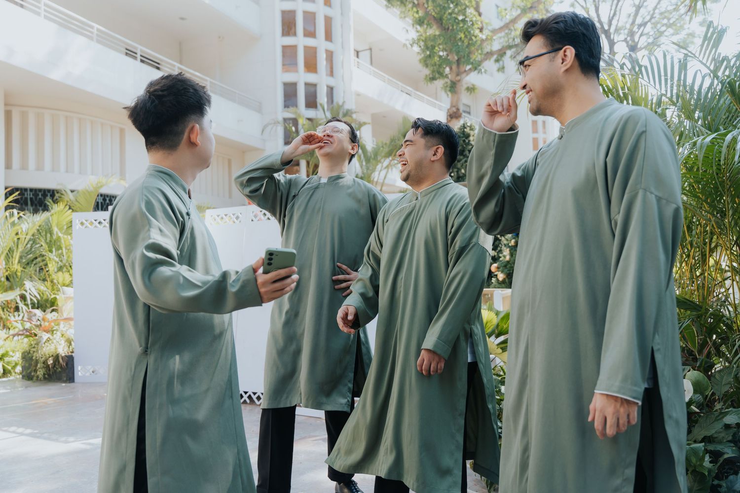 Group of men wearing matching olive green traditional outfits socializing outdoors near a white building.