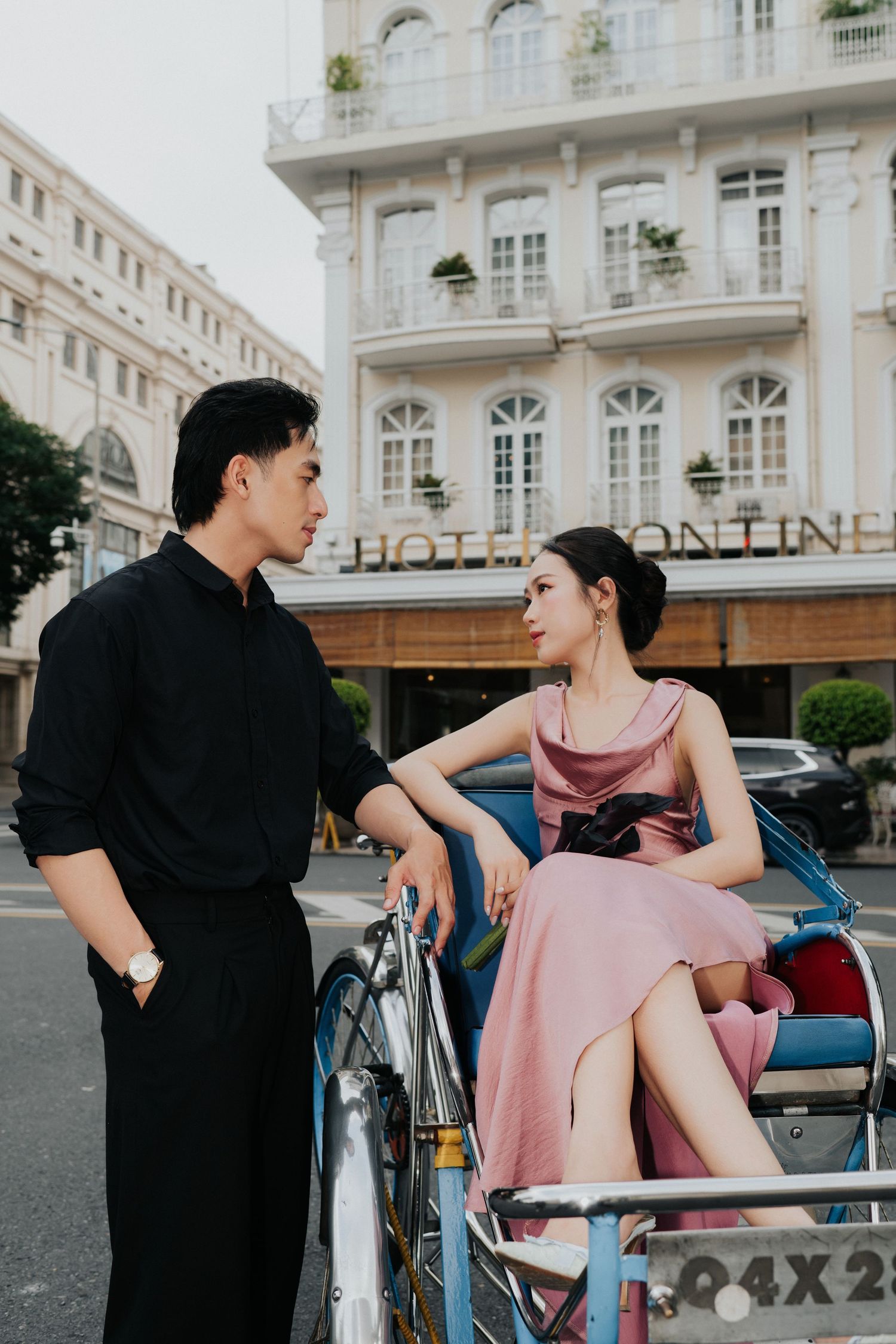 A couple in elegant attire shares a romantic moment by a rickshaw outside a classic hotel.