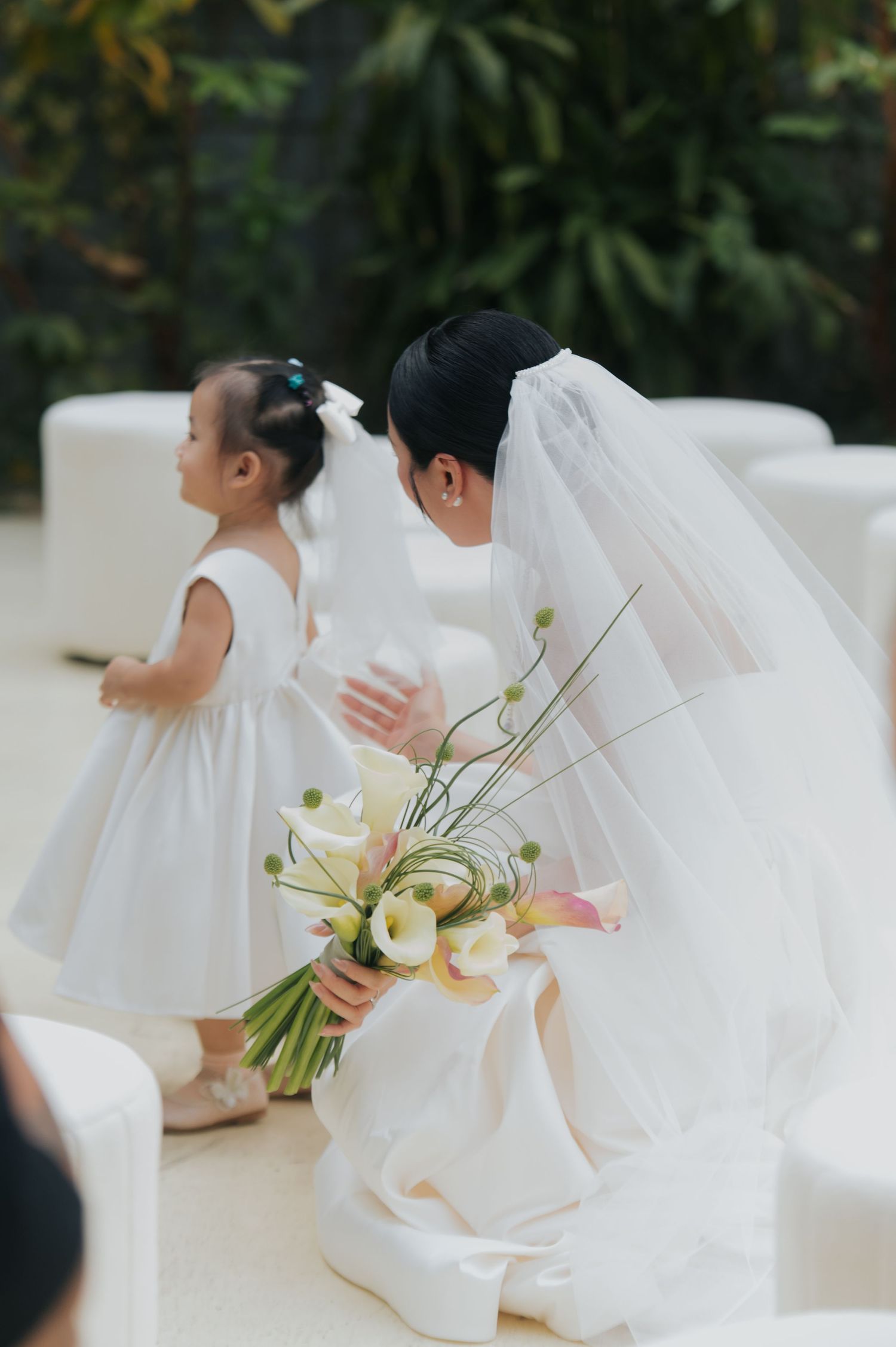 Bride in white gown and veil kneels to interact with flower girl in white dress at outdoor wedding ceremony.