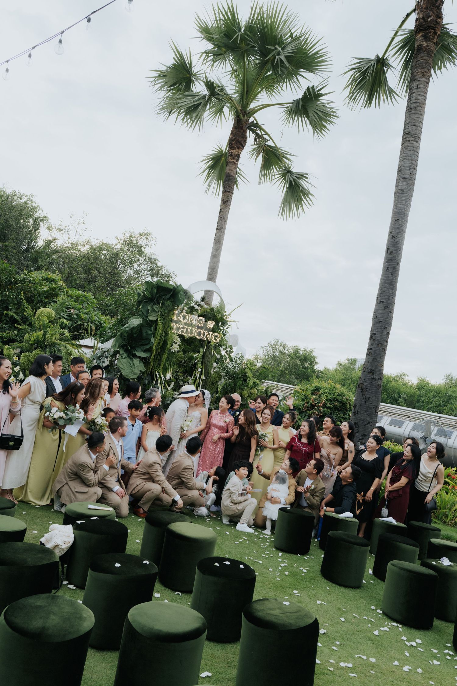 Outdoor wedding ceremony with guests seated on green velvet ottomans, palm trees, and floral arch backdrop.