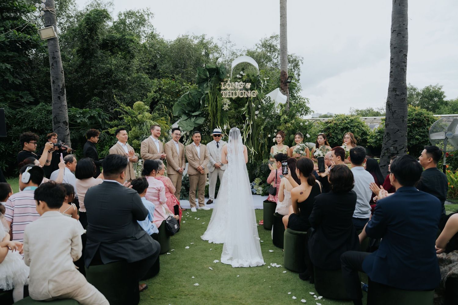 Outdoor wedding ceremony on lush green lawn with bride and groom at floral arch, surrounded by seated guests in elegant attire.