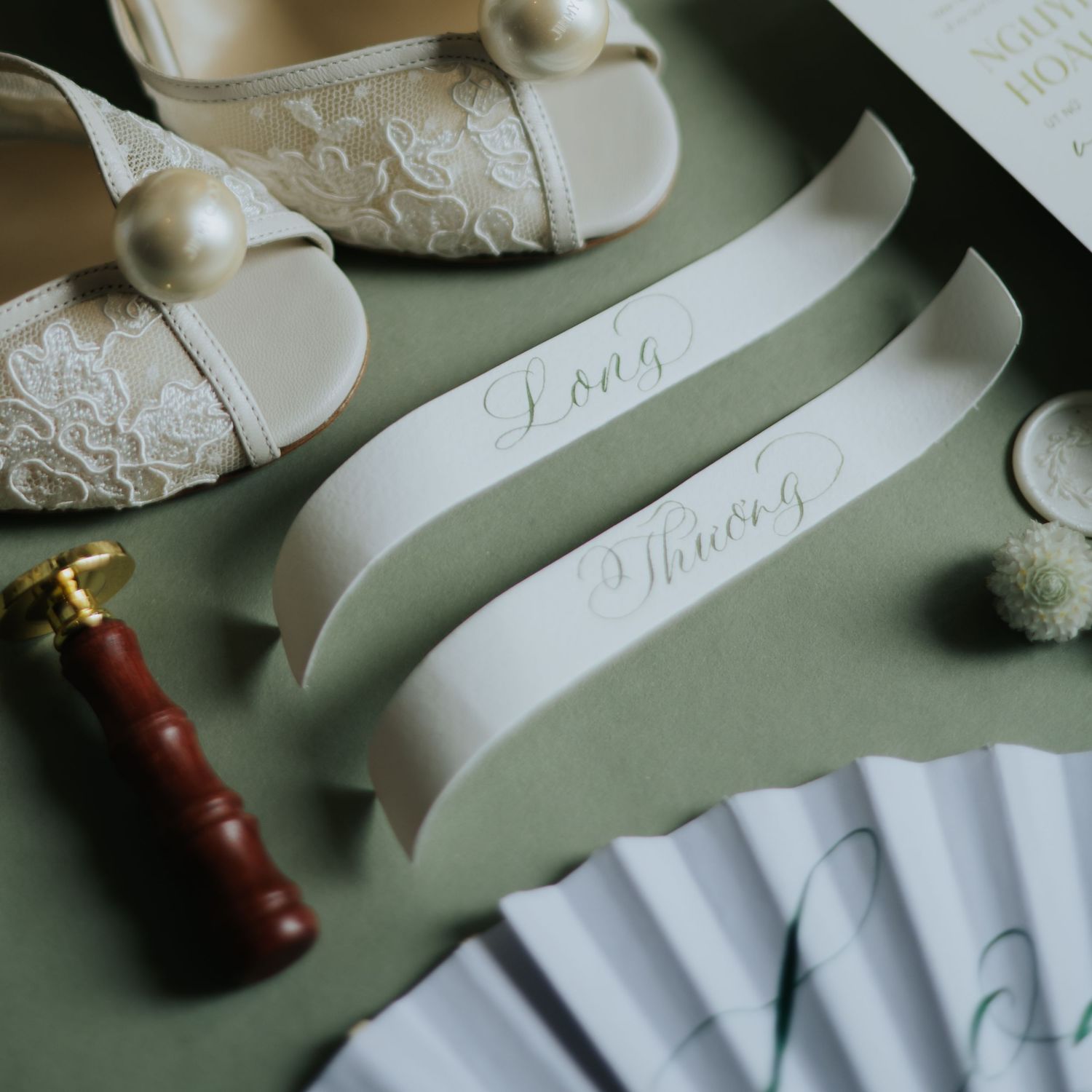 Elegant wedding flat lay with lace bridal shoes, calligraphy ribbons, wax seal, and paper fan on sage green.
