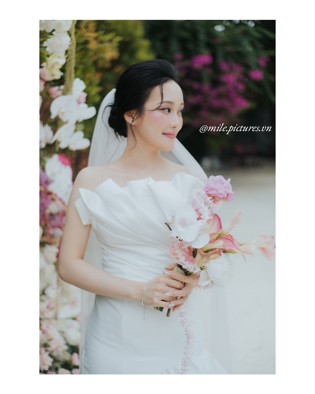Bride in white off-shoulder gown holding pink floral bouquet, wearing veil, posed near flower arch.